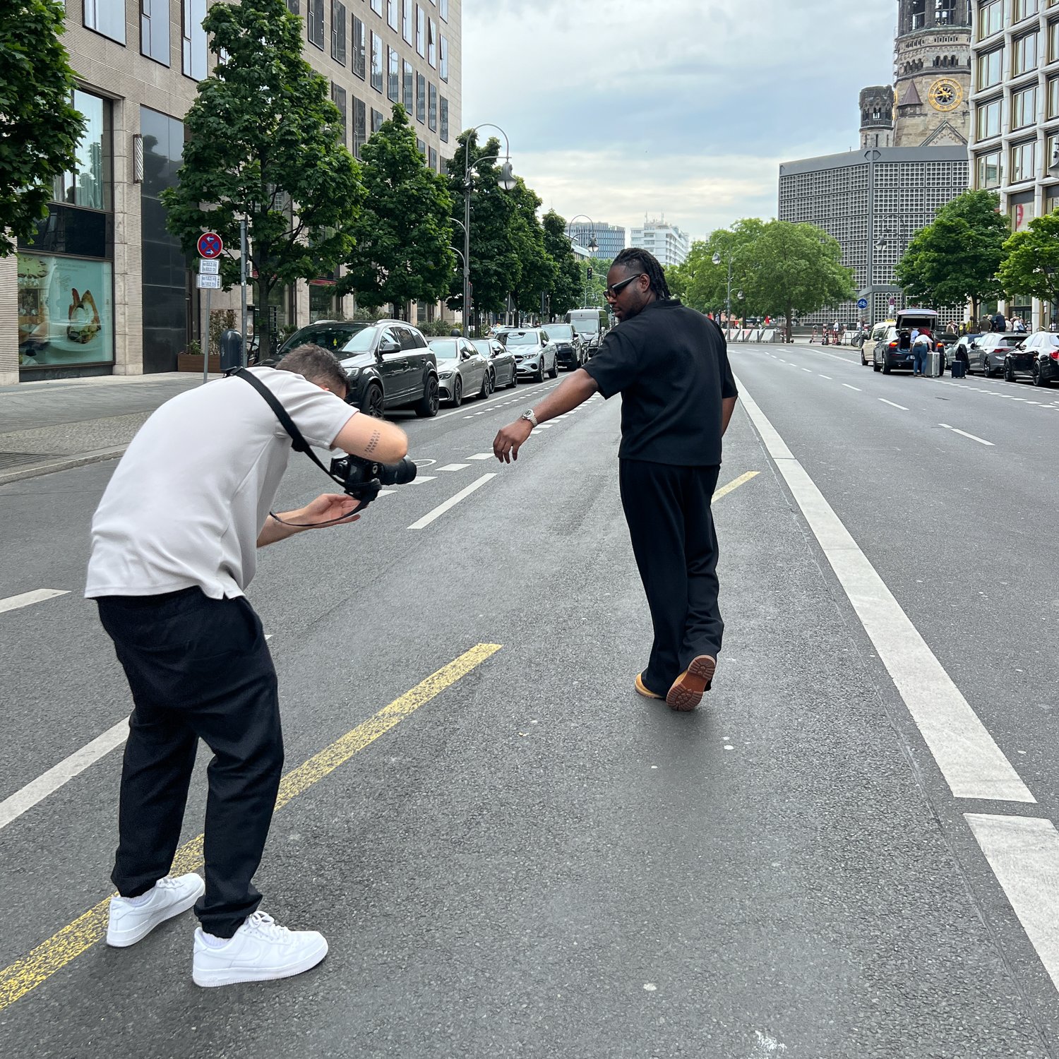 Zwei Männer auf einer Stadtstraße, einer fotografiert den anderen, der den Arm ausstreckt.