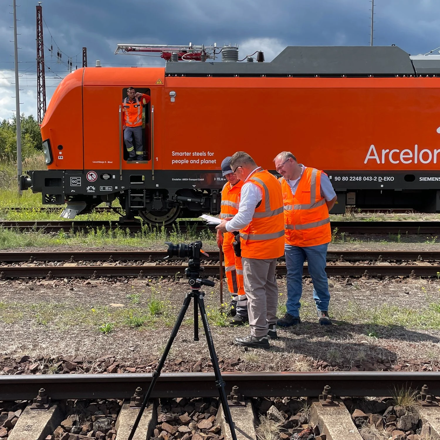 Vier Männer in orange Sicherheitswesten stehen neben Bahngleisen und prüfen Dokumente, während eine Person im Zug ist. Es gibt eine Kamera auf einem Stativ im Vordergrund und eine orange Lokomotive im Hintergrund.