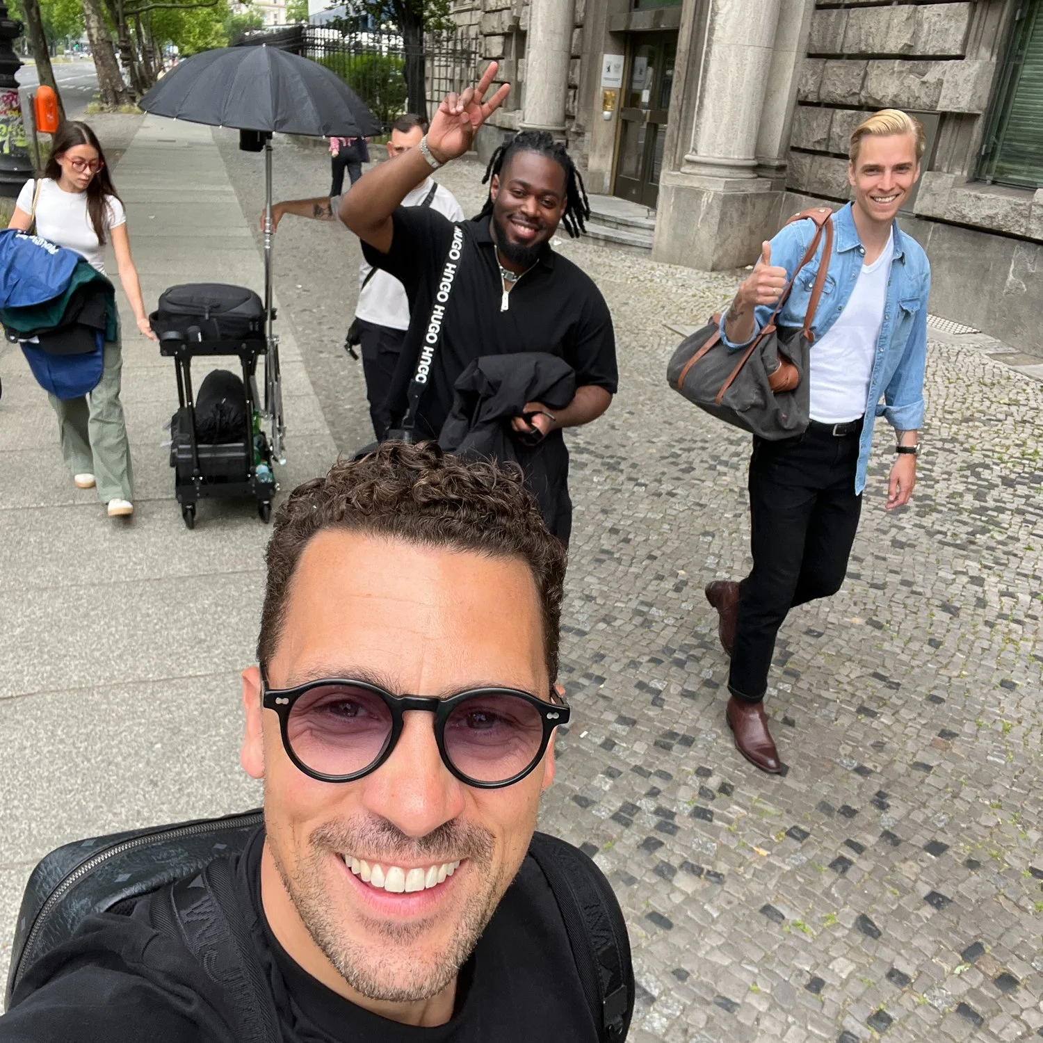 Gruppe von fünf Personen, die auf der Straße gehen, eine davon macht ein Peace-Zeichen, alle freuen sich und haben Taschen oder Rucksäcke, im Hintergrund Gebäude und Bäume, es ist bewölkt.