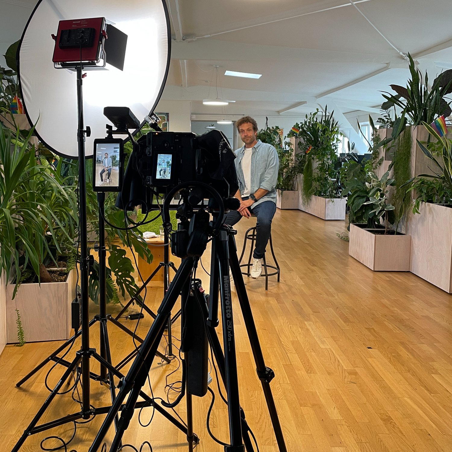 Ein Mann sitzt auf einem Barhocker vor einer Fotoleinwand, umgeben von Pflanzen in einem hellen, modernen Raum, während er für ein Interview oder eine Aufnahme posiert, mit einer Kamera und professioneller Beleuchtung im Vordergrund.