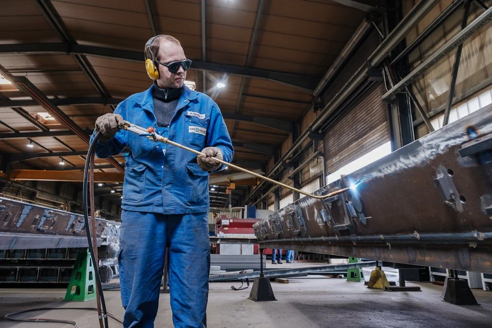 Ein Mann in blauer Arbeitskleidung schweißt an einem Metallstück in einer Werkshalle.
