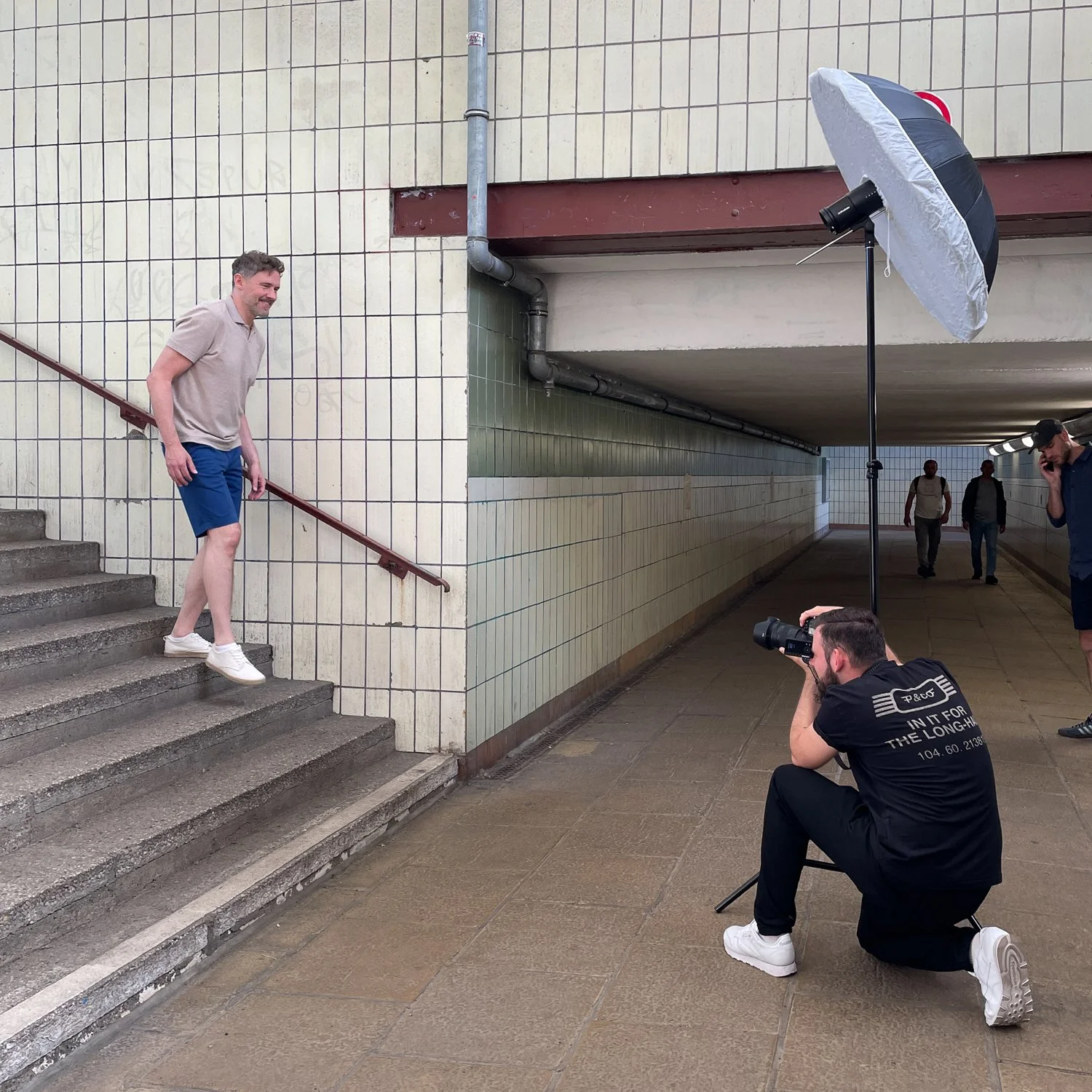 Ein Mann posiert auf den Treppen, während ein Fotograf ihn in einer Unterführung fotografiert. Es gibt auch weitere Personen im Hintergrund, die durch die Unterführung gehen.