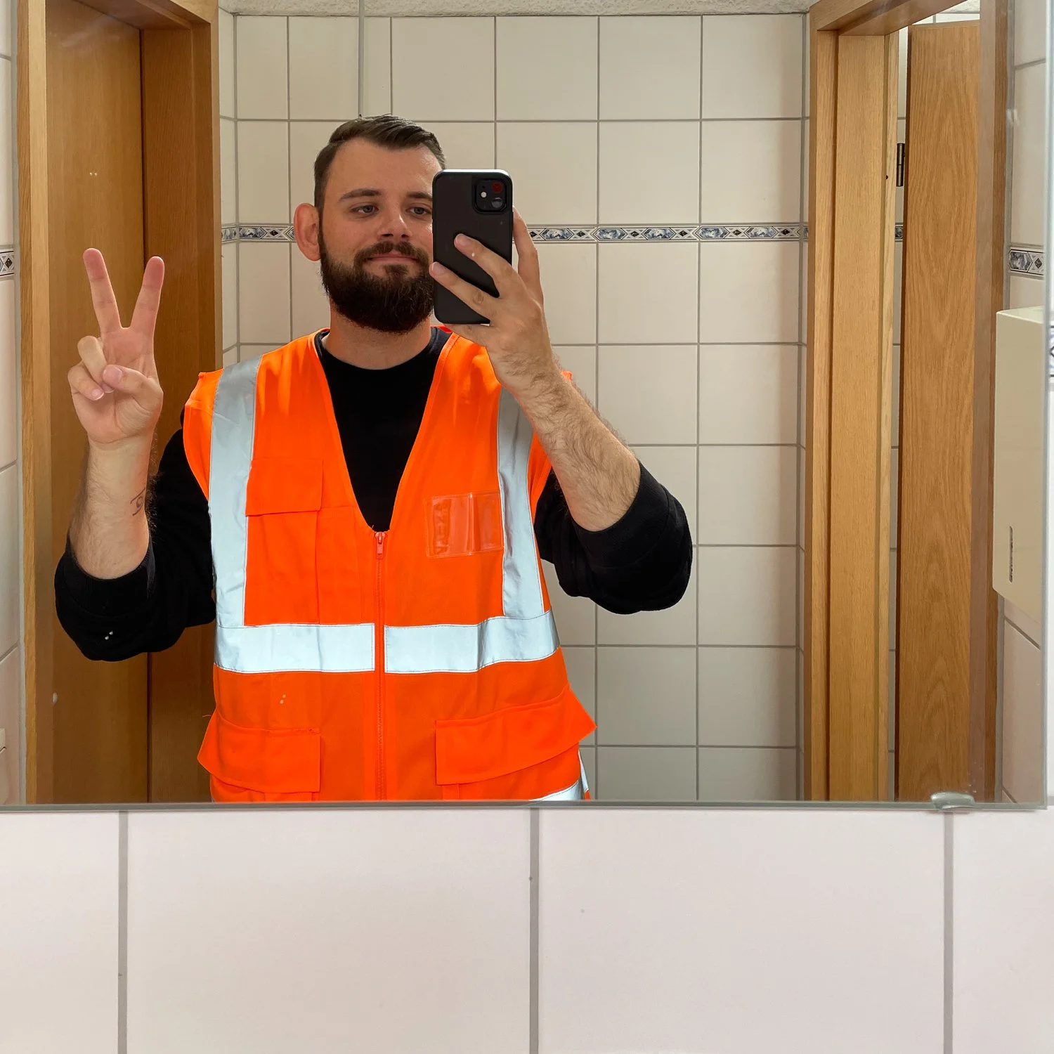 Junger Mann mit Bart trägt eine orange Warnweste und macht ein Peace-Zeichen, während er ein Selfie in einer Gut befestigten Toilette im Badezimmer macht.