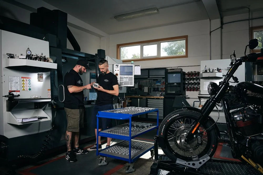 Zwei Männer in einer Werkstatt, die sich gegenseitig eine Komponente zeigen, mit einer schwarzen Harley-Davidson-Motorrad im Vordergrund und Werkzeugen im Hintergrund.