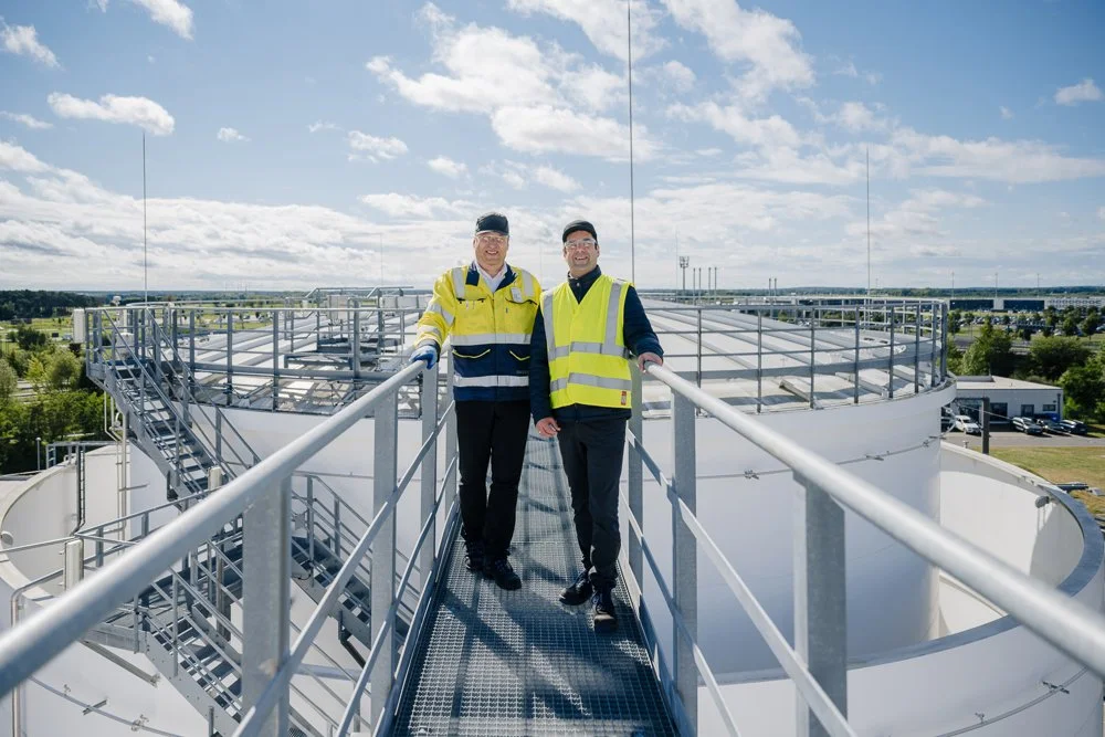 Zwei Männer in Sicherheitswesten stehen auf einer Metallbrücke auf dem Dach eines Industriegebäudes, mit einer Stadt im Hintergrund und Wolken am Himmel.
