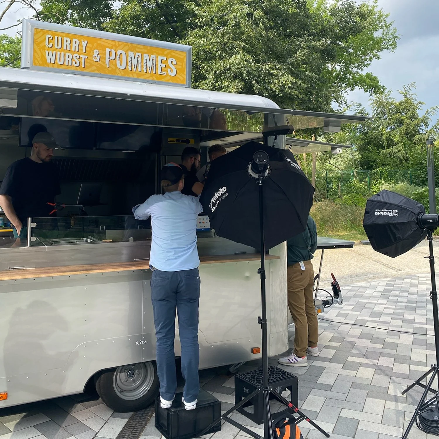 Ein Foodtruck mit einem Schild, auf dem steht 'Curry & Wurst & Pommes'. Menschen stehen vor dem Foodtruck, während Fotografie-Equipment im Einsatz ist.