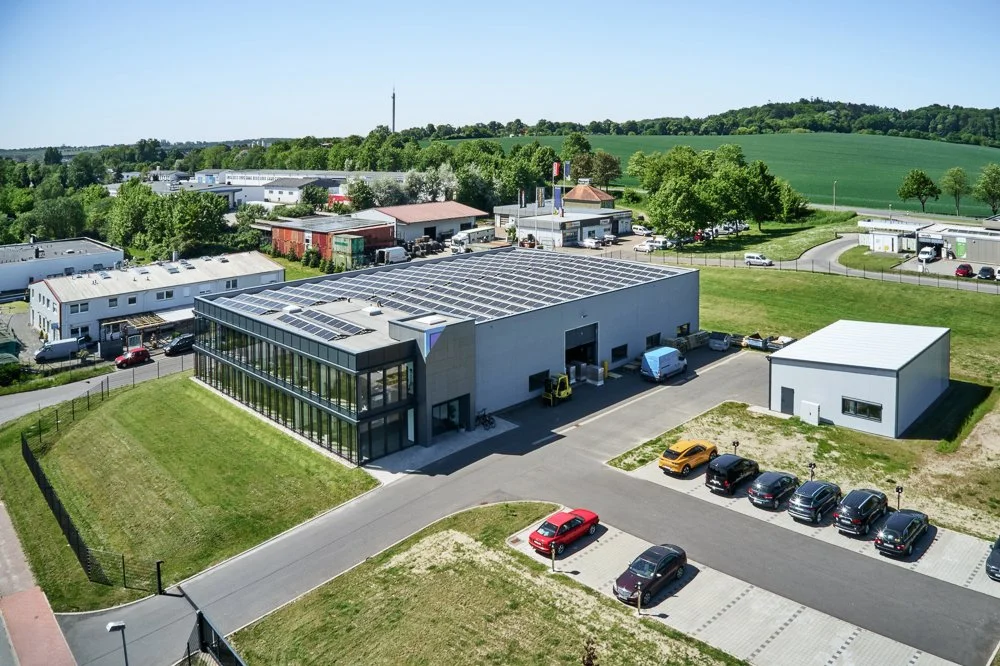 Luftaufnahme eines modernen Gebäudes mit Solarpanels auf dem Dach, umgeben von Parkplätzen und grünem Land.