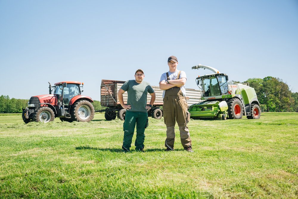 Zwei Männer stehen vor landwirtschaftlichen Maschinen auf einer grünen Wiese bei sonnigem Himmel.