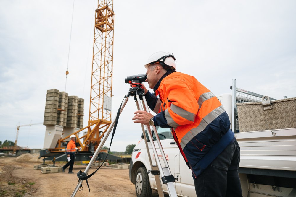 Bauarbeiter mit Schutzhelm und Jacke mit Reflektoren benutzt ein Vermessungstool auf einer Baustelle, im Hintergrund ein Kran und einen weiteren Arbeiter.