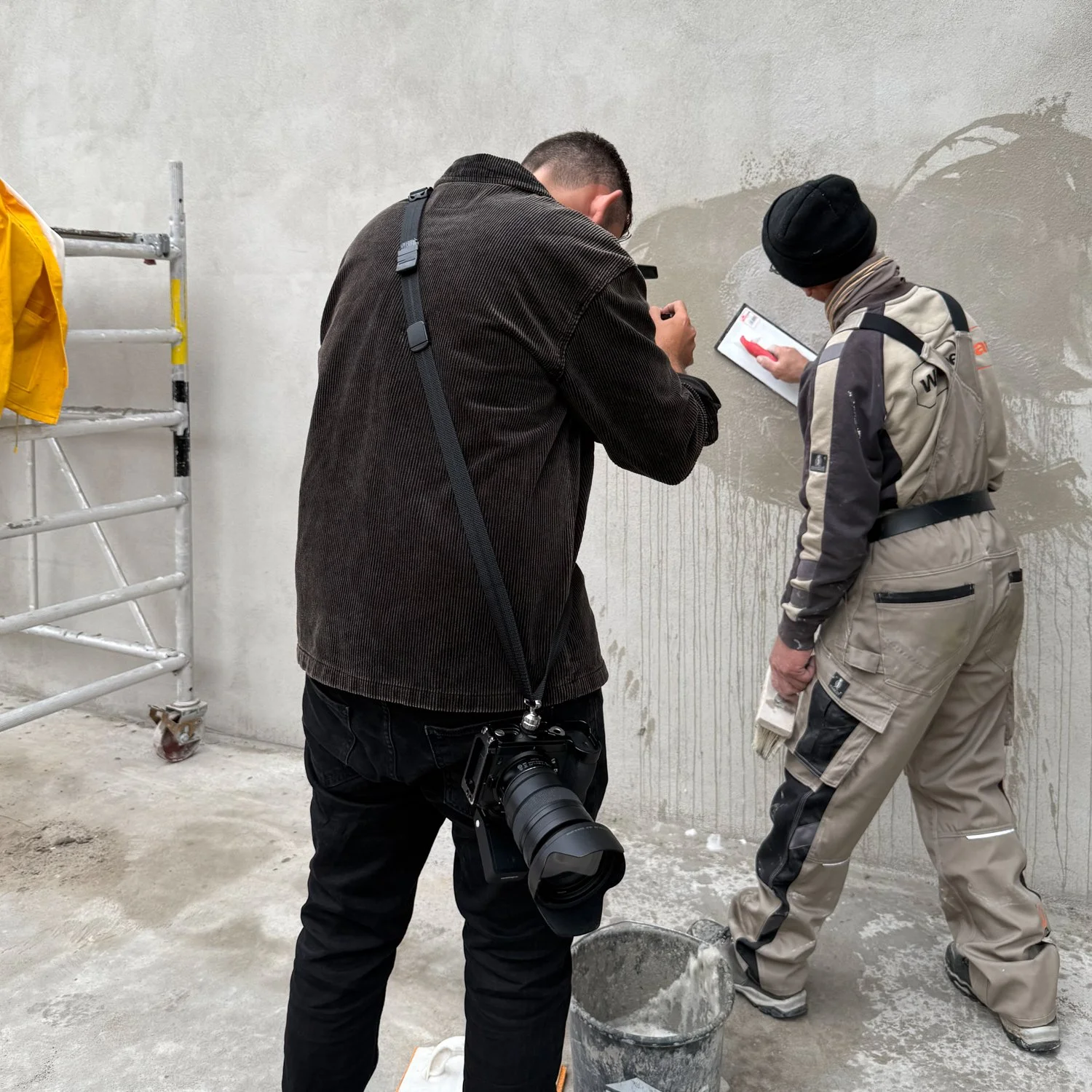 Zwei Bauarbeiter bei der Arbeit an einer Wand, während ein Fotograf Fotos macht.