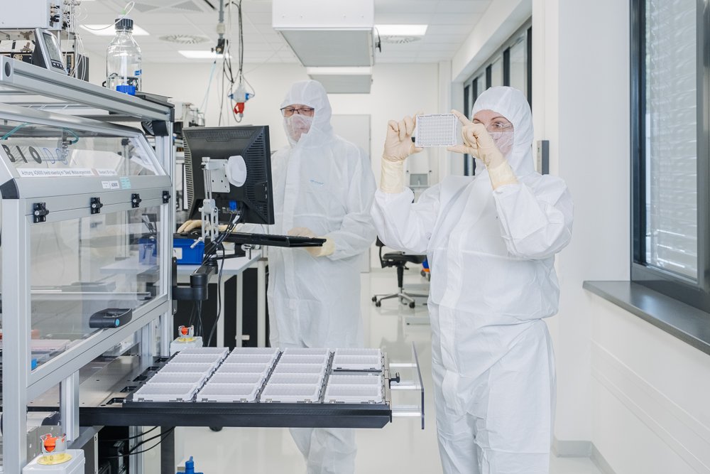 Zwei Wissenschaftler in Schutzkleidung in einem Labor, einer nimmt ein Foto von einem Probenraster auf, der andere arbeitet an einem Computer.