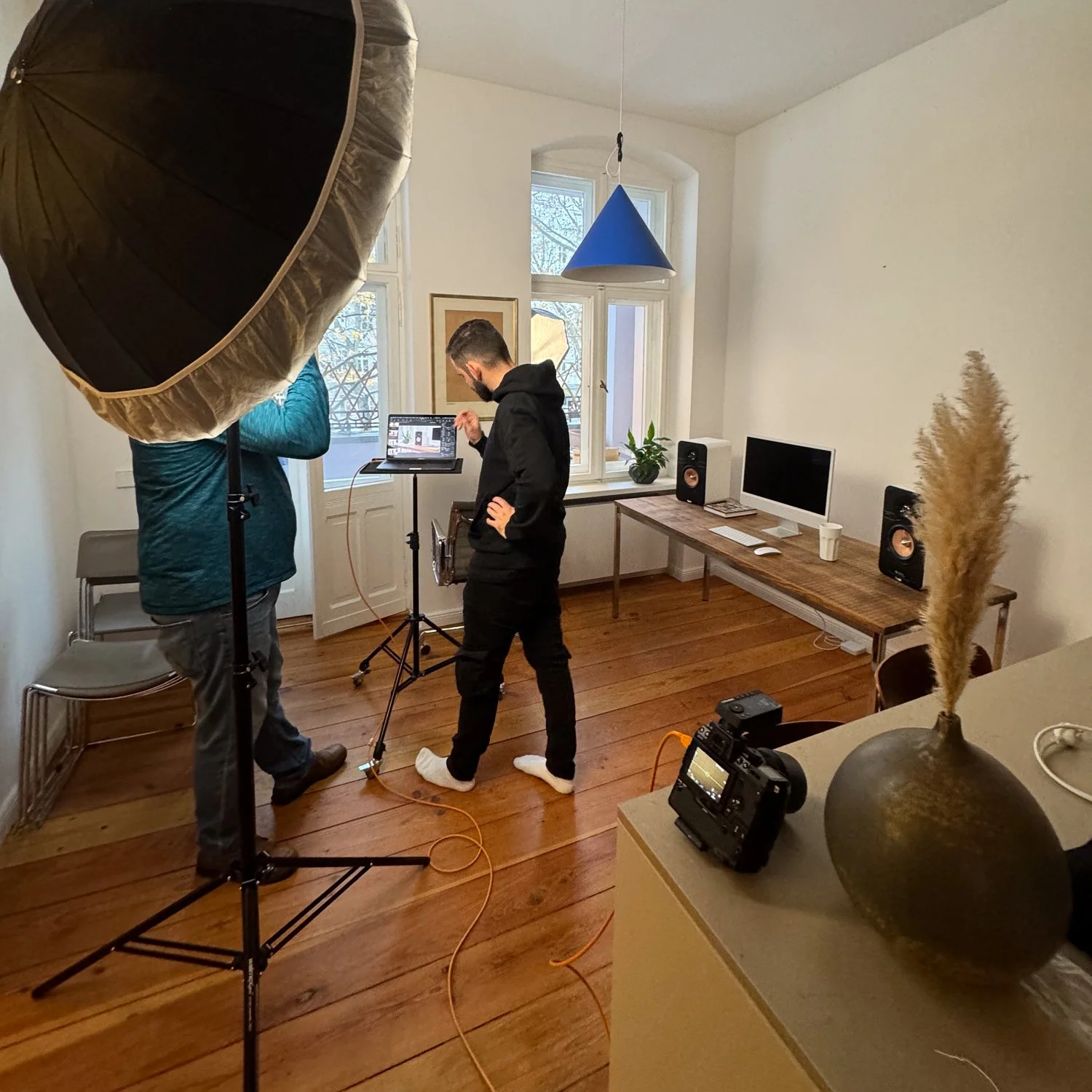 Innenraum eines Raums mit zwei Personen bei einer Fotosession. Es gibt Beleuchtungszubehör, einen Laptop, einen Desktop-Computer, Lautsprecher auf einem Tisch, eine Blumenvase mit Trockenblumen und eine Kamera auf einem Tisch im Vordergrund.