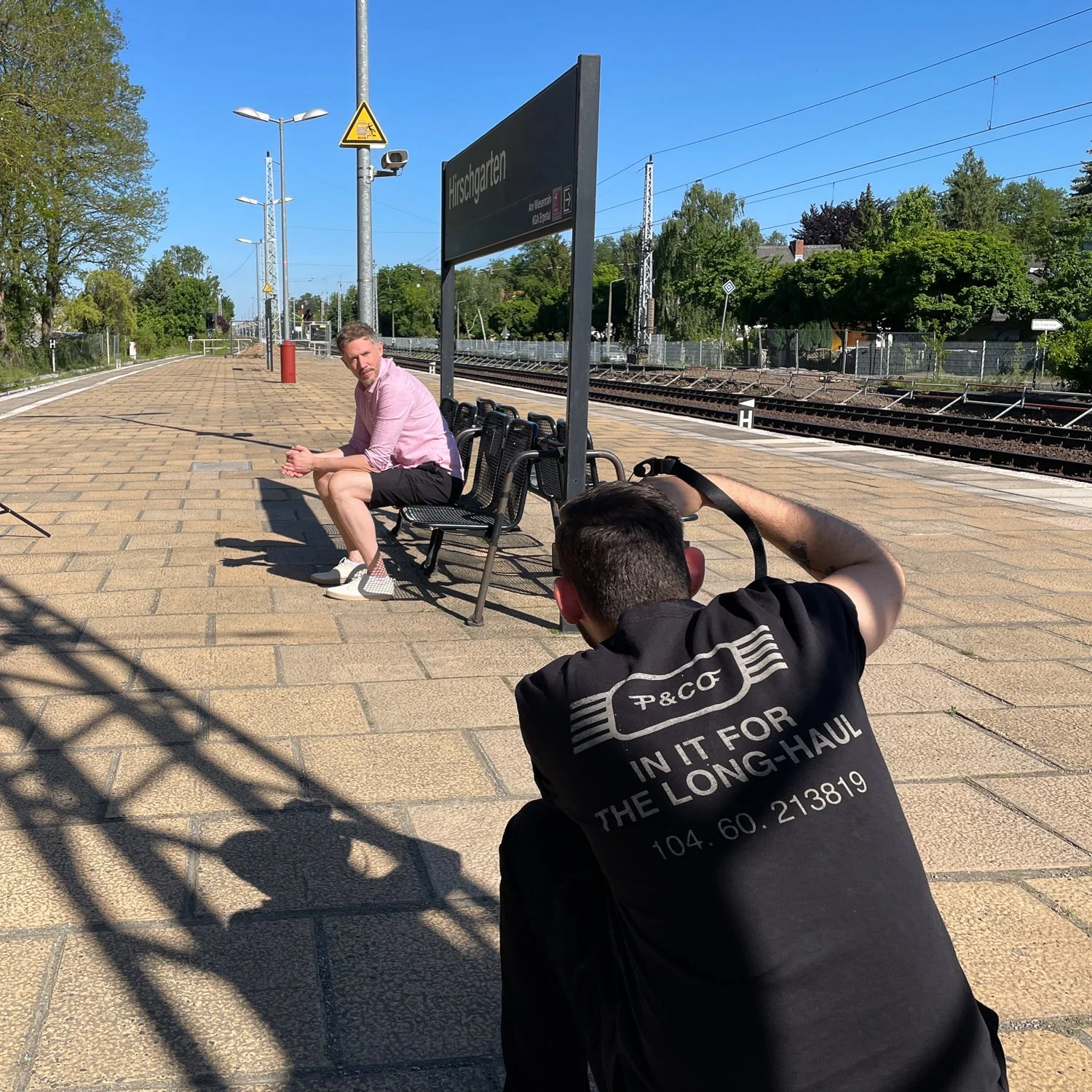 Ein Fotograf macht ein Bild von einem Mann auf einem Bahnsteig bei Sonnenschein, im Hintergrund sind Gleise und Bäume, der Mann sitzt auf einer Bank und trägt ein pinkes Hemd und schwarze Shorts.