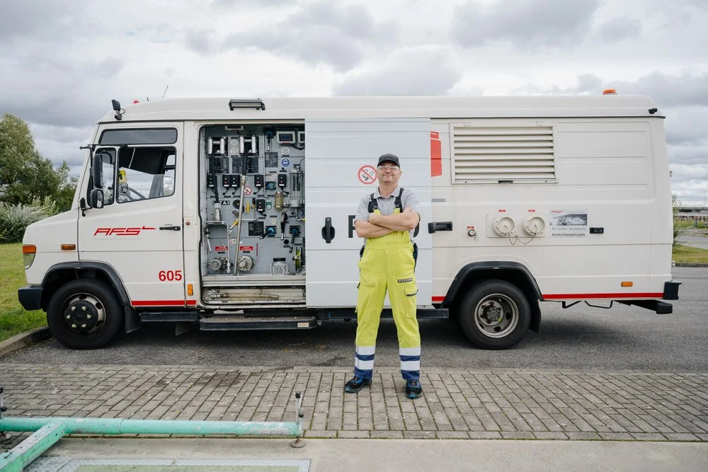 Feuerwehrmann in Warnkleidung steht vor einem weißem Feuerwehrauto mit offenem Seitenorganisationsfach und technischen Geräten.