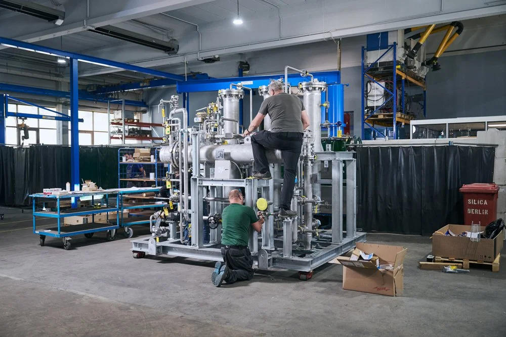 Zwei Männer arbeiten an industrieller Maschine in einer Fabrikhalle mit Werkzeugwagen und Kartons.