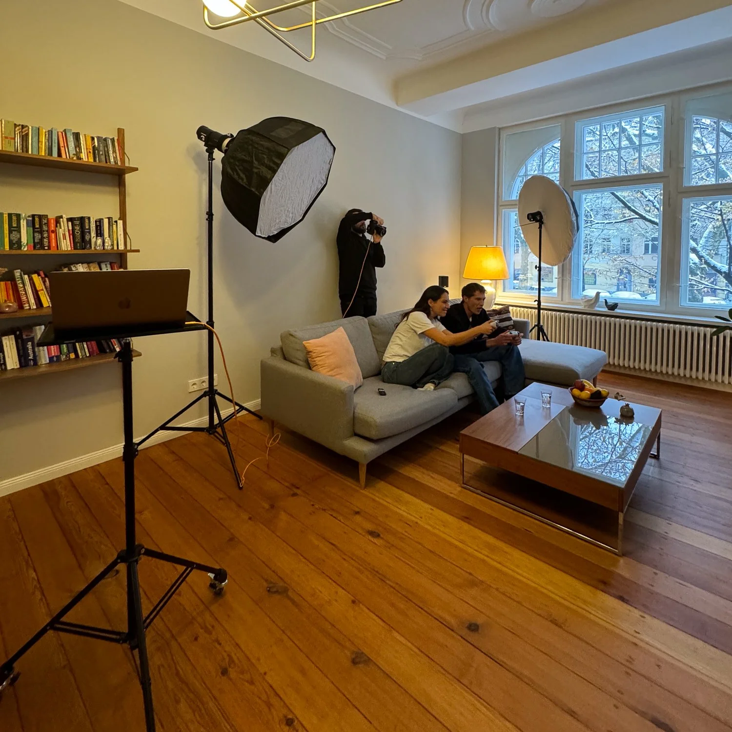 Zwei Personen sitzen auf einem grauen Sofa, während eine Person im Hintergrund fotografiert wird, mit professioneller Fotoausrüstung und Licht. Das Wohnzimmer hat große Fenster, einen Couchtisch mit Obst und Gläsern, sowie Bücherregale.