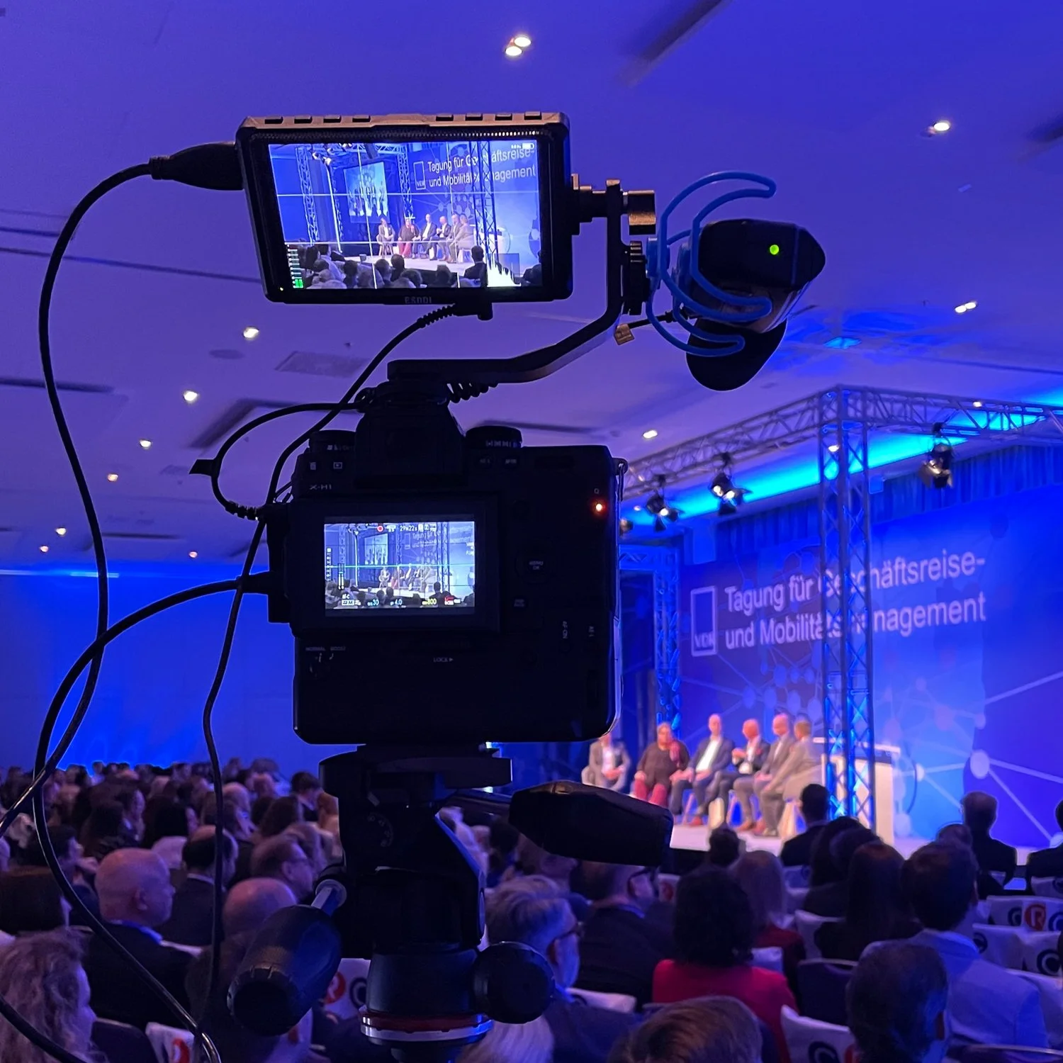 Aufnahme einer Konferenz mit mehreren Sprechern auf der Bühne, aufgenommen durch eine Kamera mit Blick auf das Publikum, das in einem großen Saal sitzt.