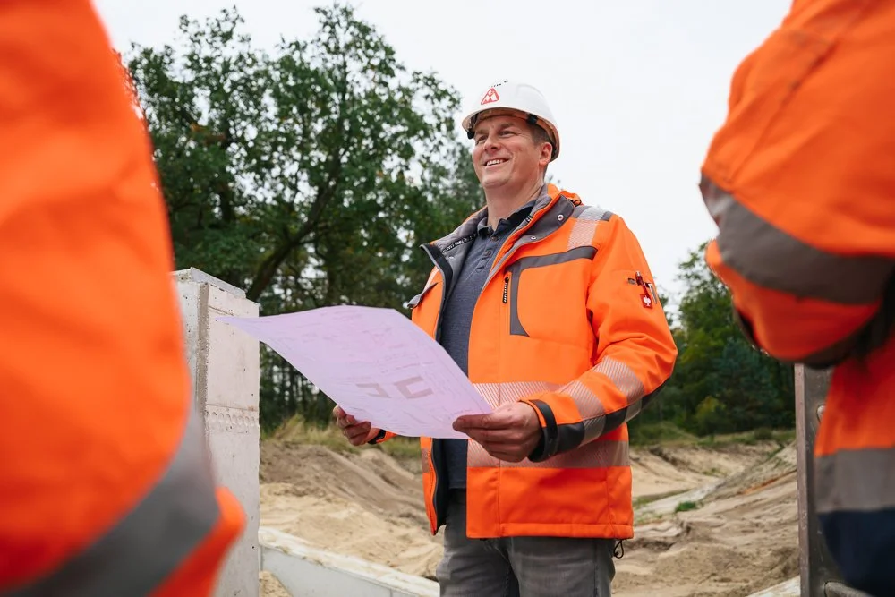 Ein Mann in orange Sicherheitsjacke und Helm steht auf einer Baustelle, hält Baupläne in der Hand und lächelt.