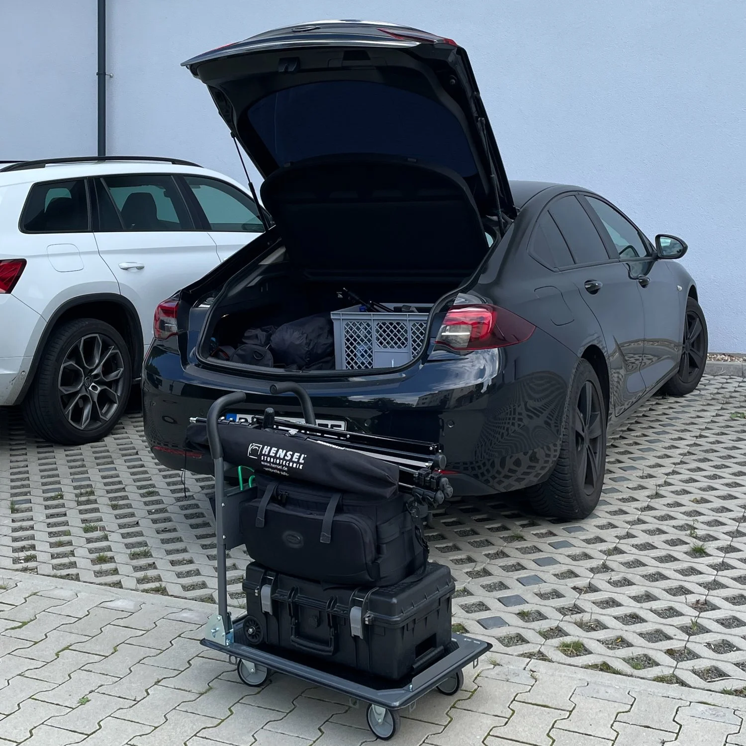 Ein schwarzer Sportwagen mit offengemachten Kofferraum, daneben ein Gepäckwagen mit Kameraausrüstung und anderen Gepäckstücken. Es ist ein zweiter weißer Auto im Hintergrund.