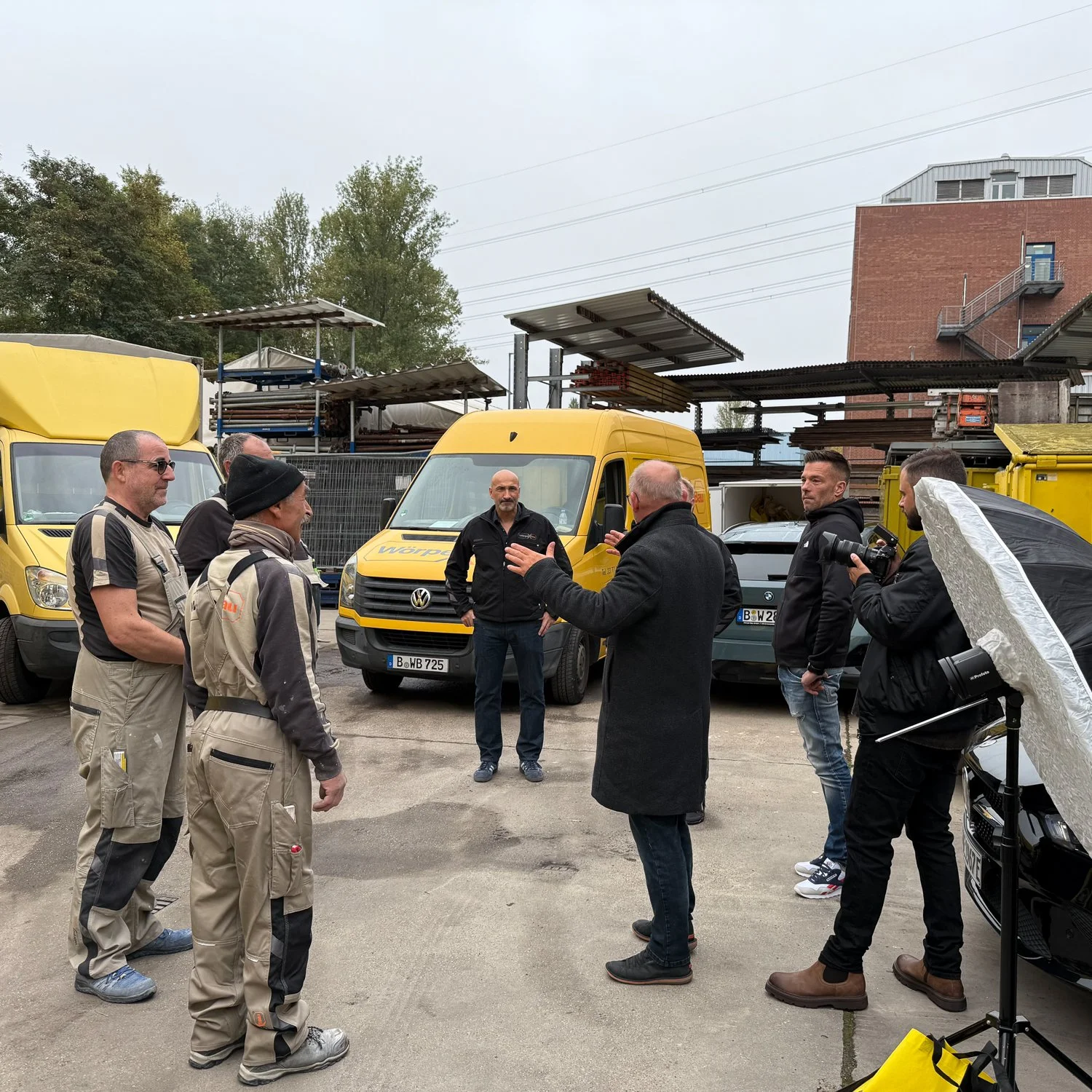 Eine Gruppe von Menschen im Gespräch in einer Auto-Werkstatt oder einem Lager mit mehreren gelben Lieferwagen im Hintergrund.