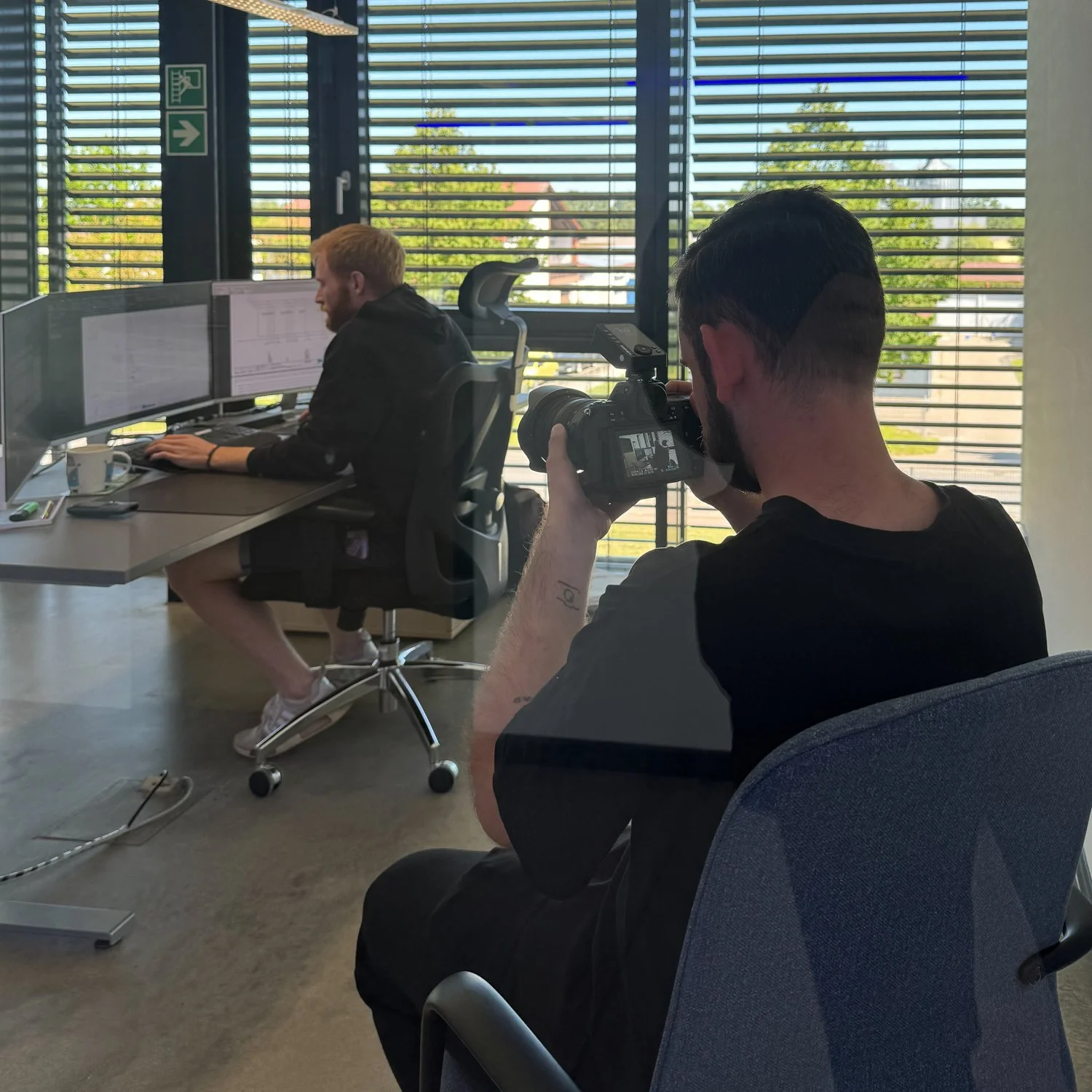 Ein Mann fotografiert eine andere Person im Büro mit großen Monitoren und einer Fensterfront im Hintergrund.