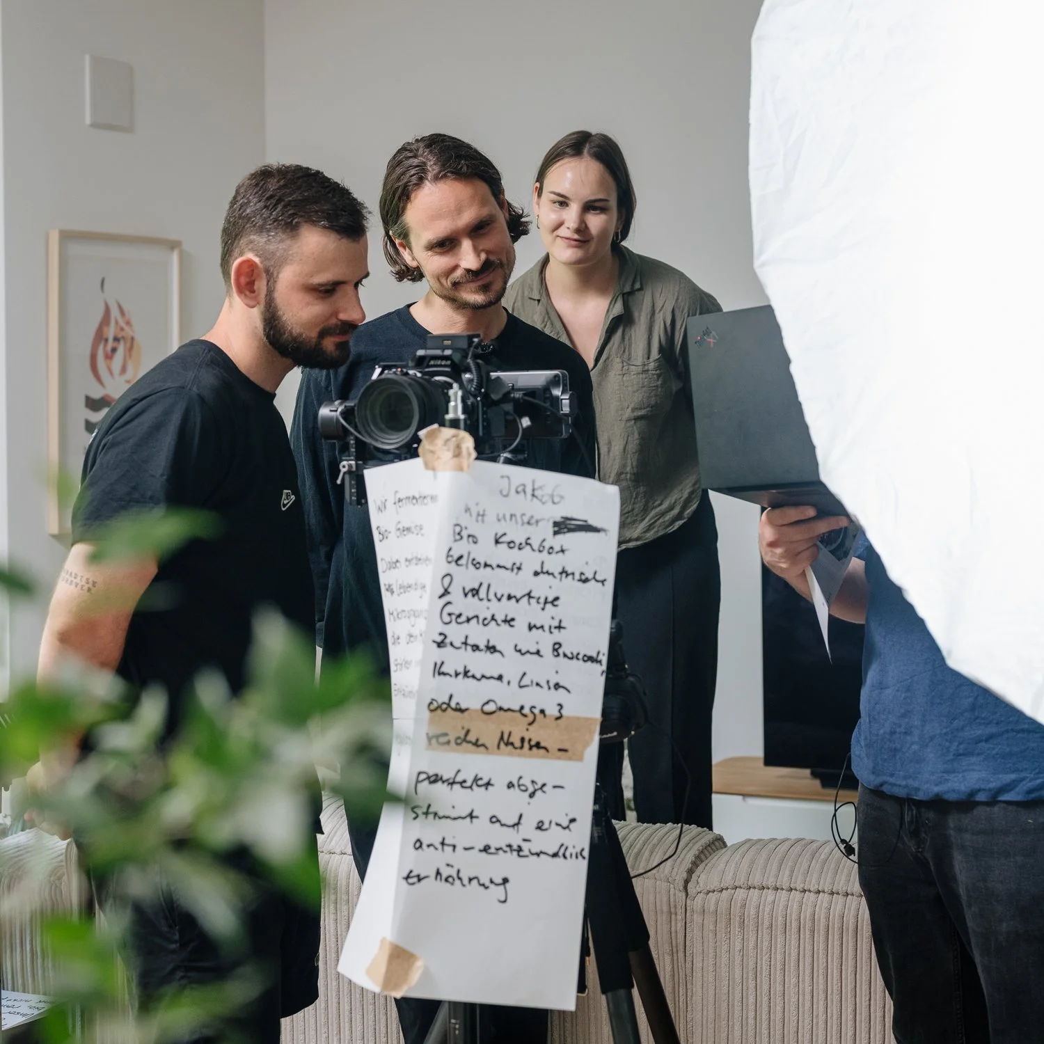 Eine Gruppe von drei Menschen beobachtet eine Kamera auf einem Stativ, mit einem großen Whiteboard oder Notizzettel mit handgeschriebenen Notizen. Sie befinden sich in einem modernen Wohnzimmer mit einer Couch und Kunstwerken an der Wand. Es sieht na