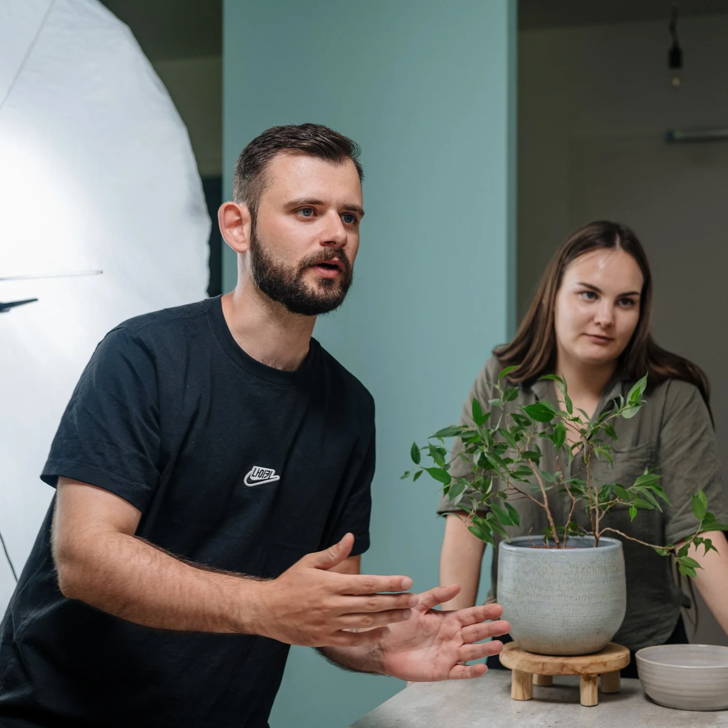 Zwei Personen, ein Mann mit dunklem T-Shirt und eine Frau mit grünem Hemd, sprechen bei einer Plattform für Pflanzen, im Vordergrund eine Topfpflanze auf einem Tablett.