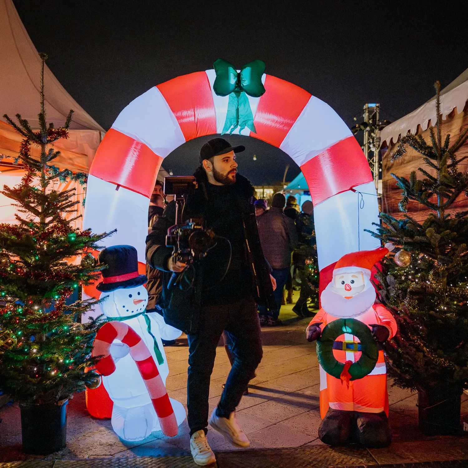 Ein Mann mit dunklem Hut und schwarzen Kleidungsstücken, der eine Kamera trägt, steht vor einer Weihnachtsdekoration mit beleuchteter Schneemann- und Santa-Figur. Hintergrund mit weiteren Weihnachtsart decorations und Menschen auf einem festlichen Ma