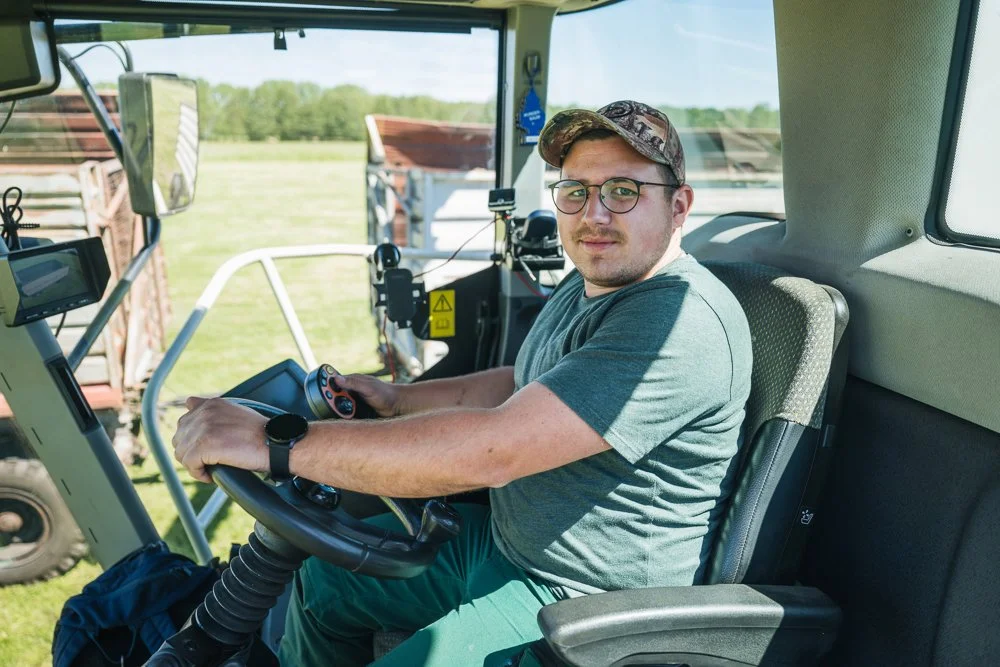 Ein junger Mann sitzt im Steuerhaus eines Traktors, trägt eine Brille, eine Mütze und ein graues T-Shirt, und hält das Lenkrad während der Fahrt auf einem Feld, im Hintergrund sind Grünsland und weitere landwirtschaftliche Fahrzeuge sichtbar.