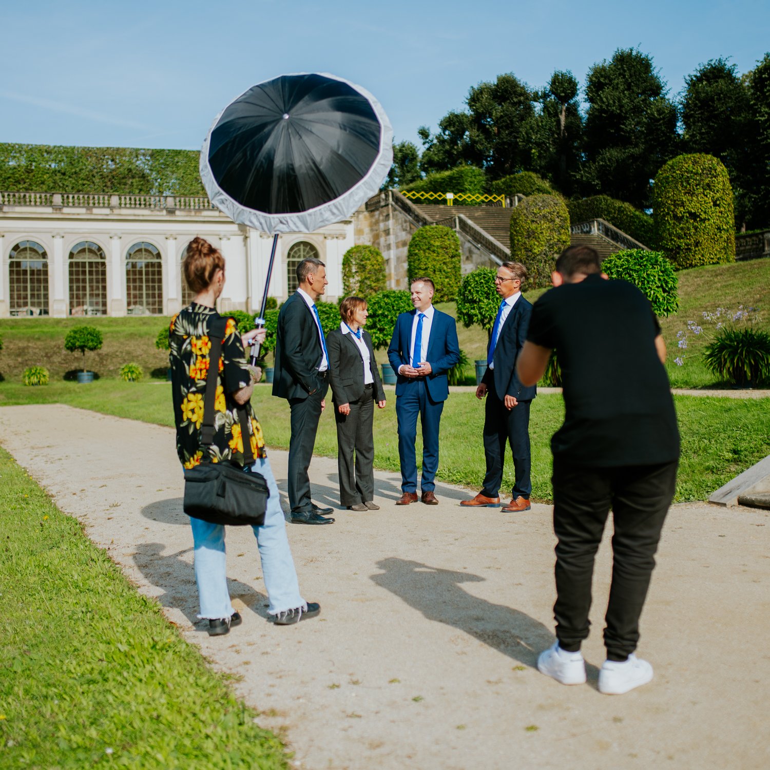 Fotoshooting im Garten mit fünf Personen, einer davon mit einem Regenschirm, die Sommerkleidung tragen, bei Sonnenschein.