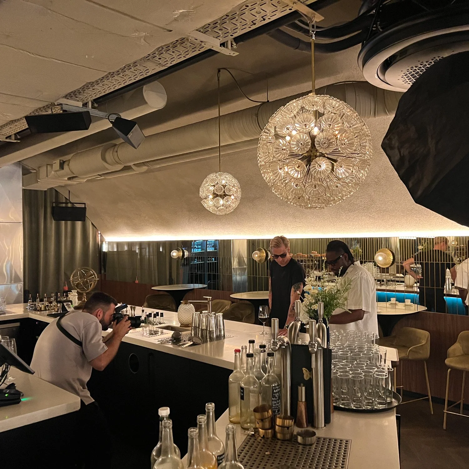 Fotografie eines modernen Bars mit zwei Männern, die Getränke zubereiten, und einem Fotografen, der Bilder macht. Das Innendesign ist stilvoll mit eleganten Lichtern und Dekorationen.