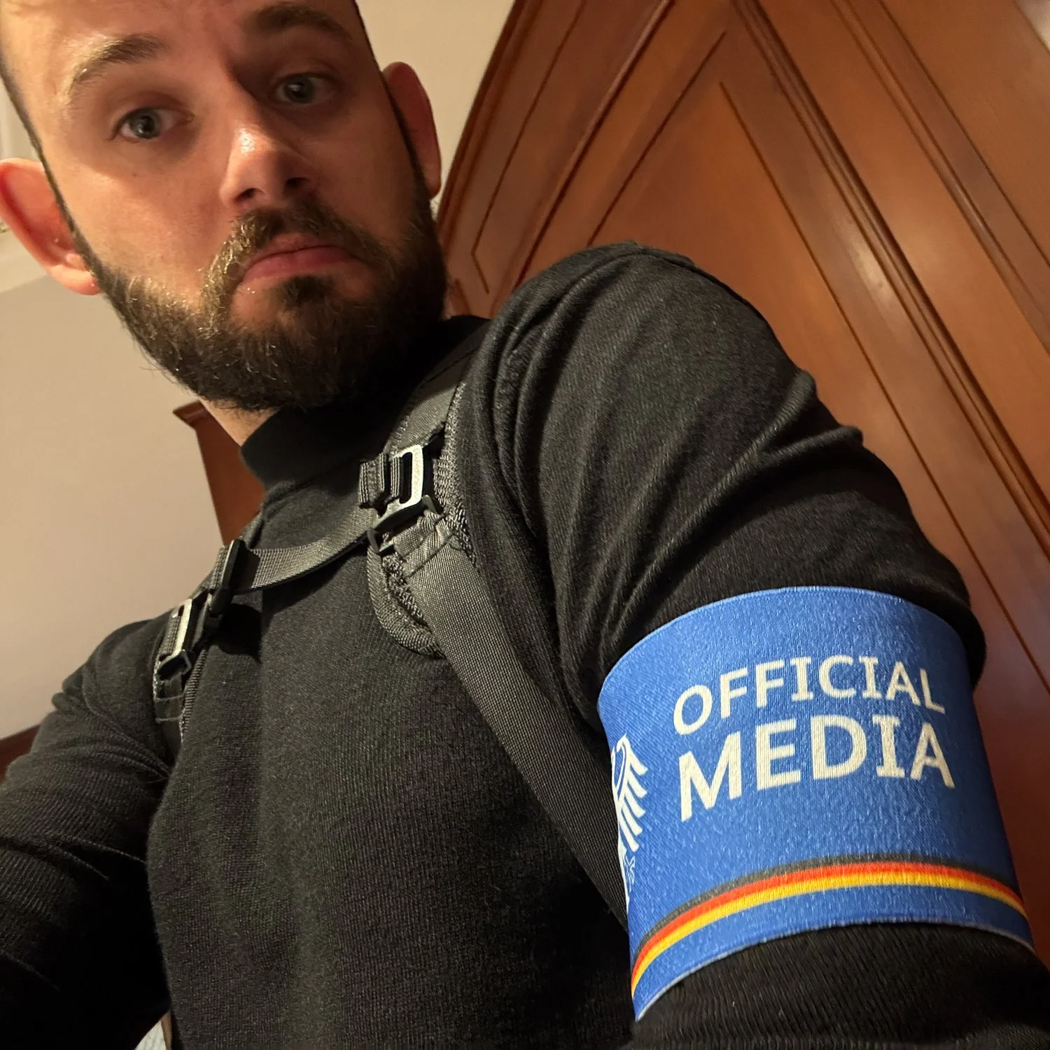 Ein Mann trägt eine schwarze Jacke und einen Rucksack, mit einem blauen Armband, auf dem 'Official Media' steht.