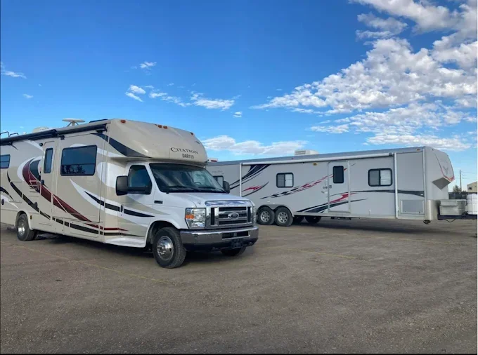 Great West Storage - A white Ford motorhome and a large white travel trailer with red and black stripes parked on a dirt lot under a partly cloudy sky.