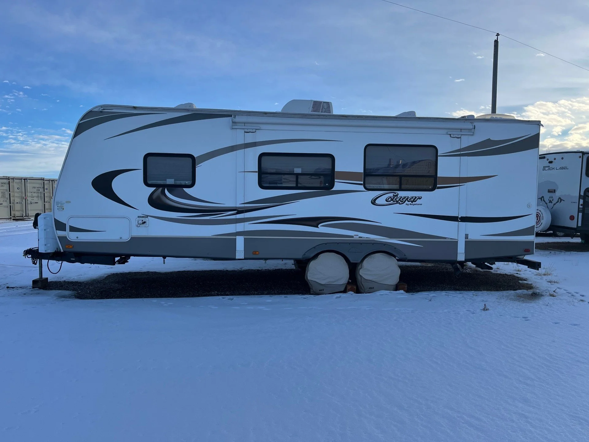 Great West Storage - White travel trailer with black and brown curved stripe graphics, parked on snow-covered ground, under a blue sky with some clouds.