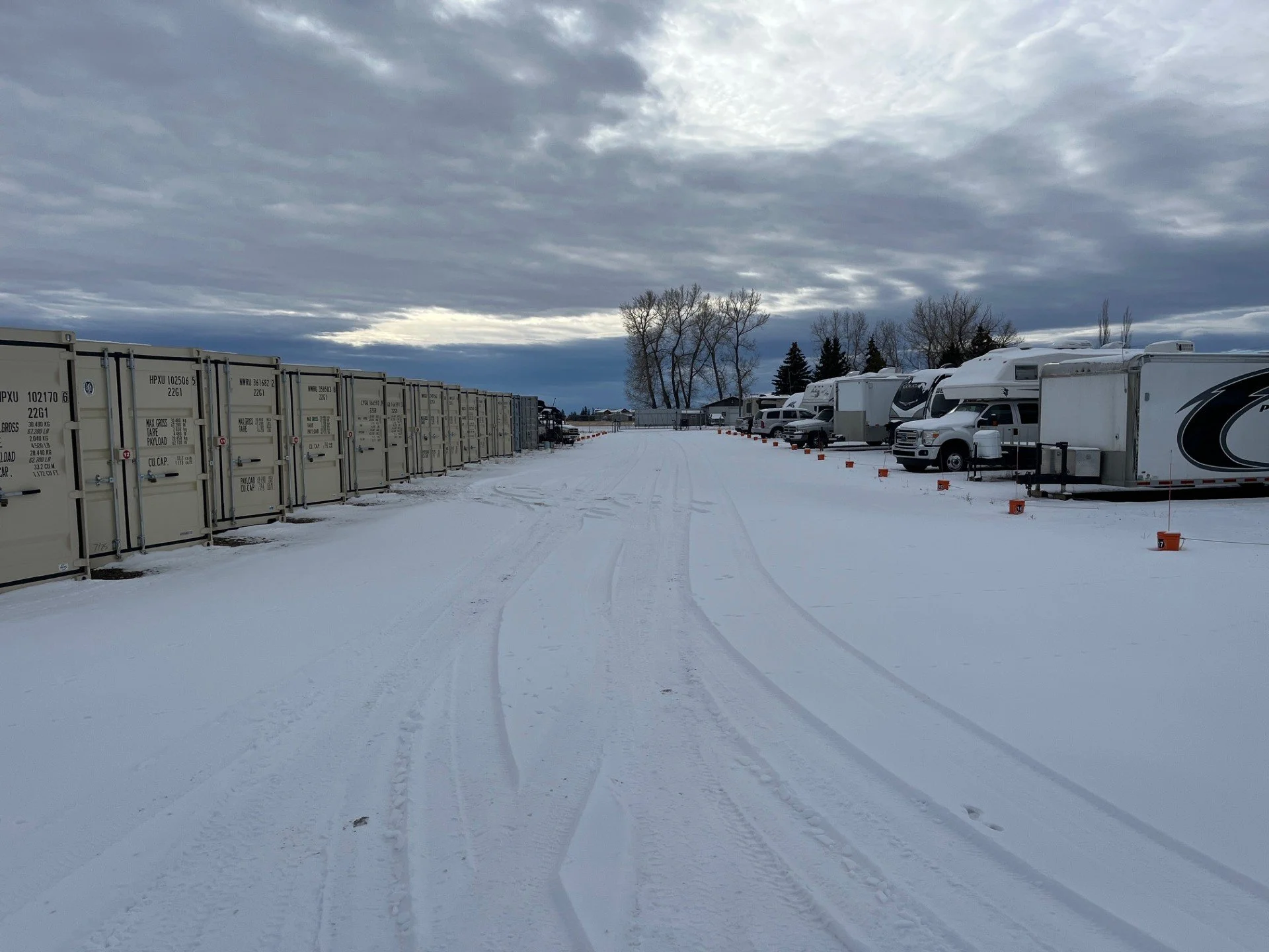 Snow-covered parking lot with travel trailers on the right and row of storage containers on the left, under a cloudy sky.