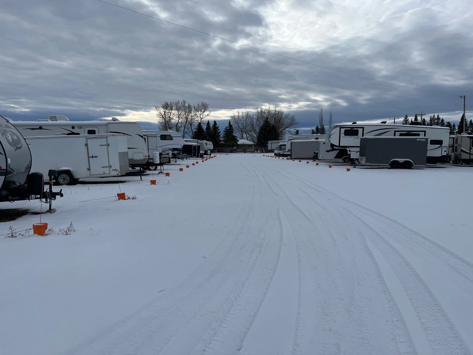 Great West Storage - Snow-covered RV parking lot with multiple RVs and trailers parked on either side, tire tracks in the snow, and a cloudy sky overhead.