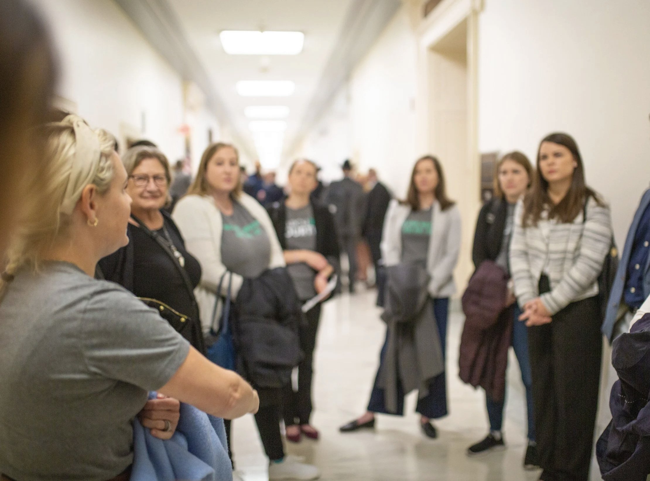 Group of people standing in a hallway, listening to a woman who is speaking, some holding coats and bags.