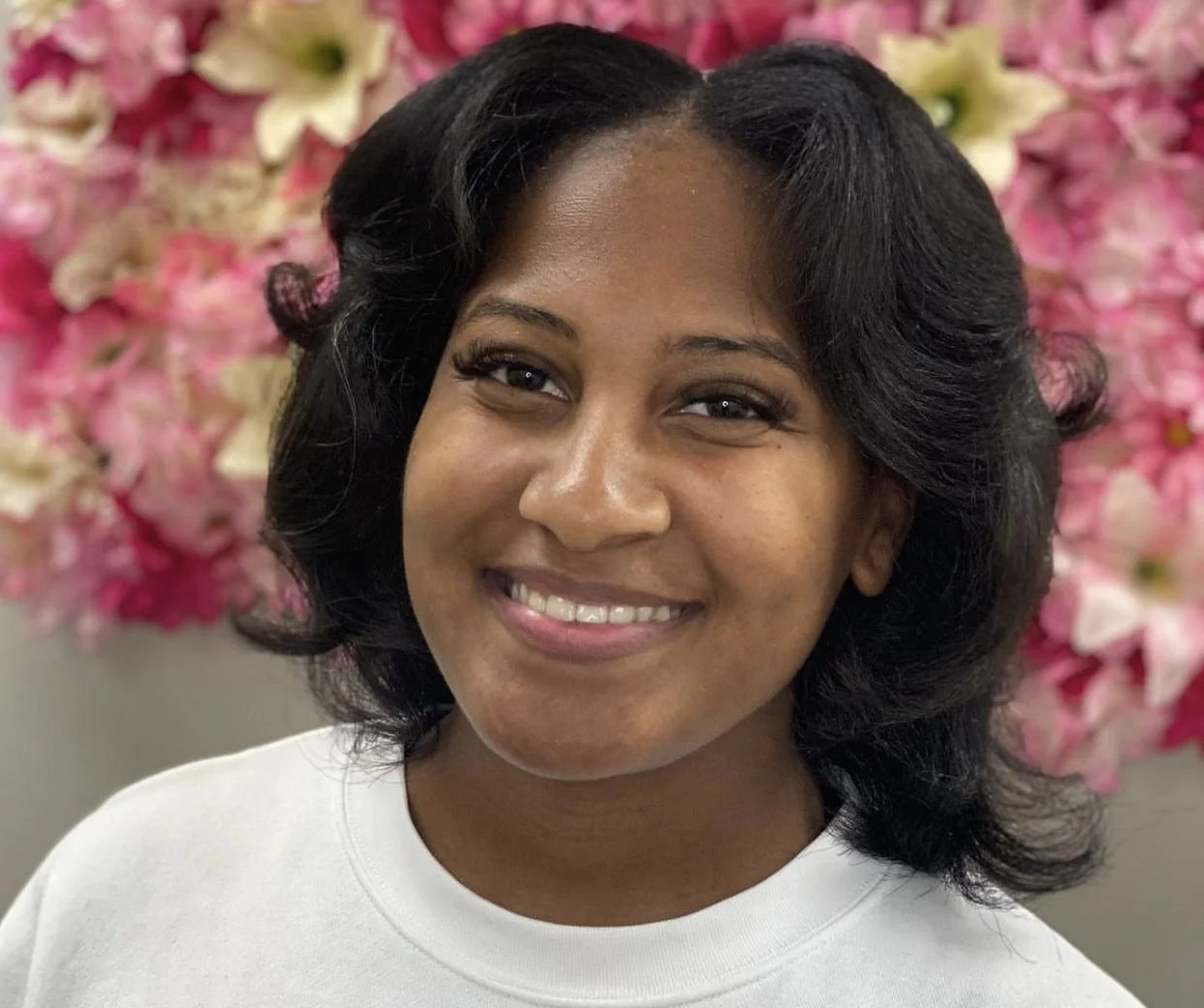 A person smiling in front of a backdrop of pink and white flowers.