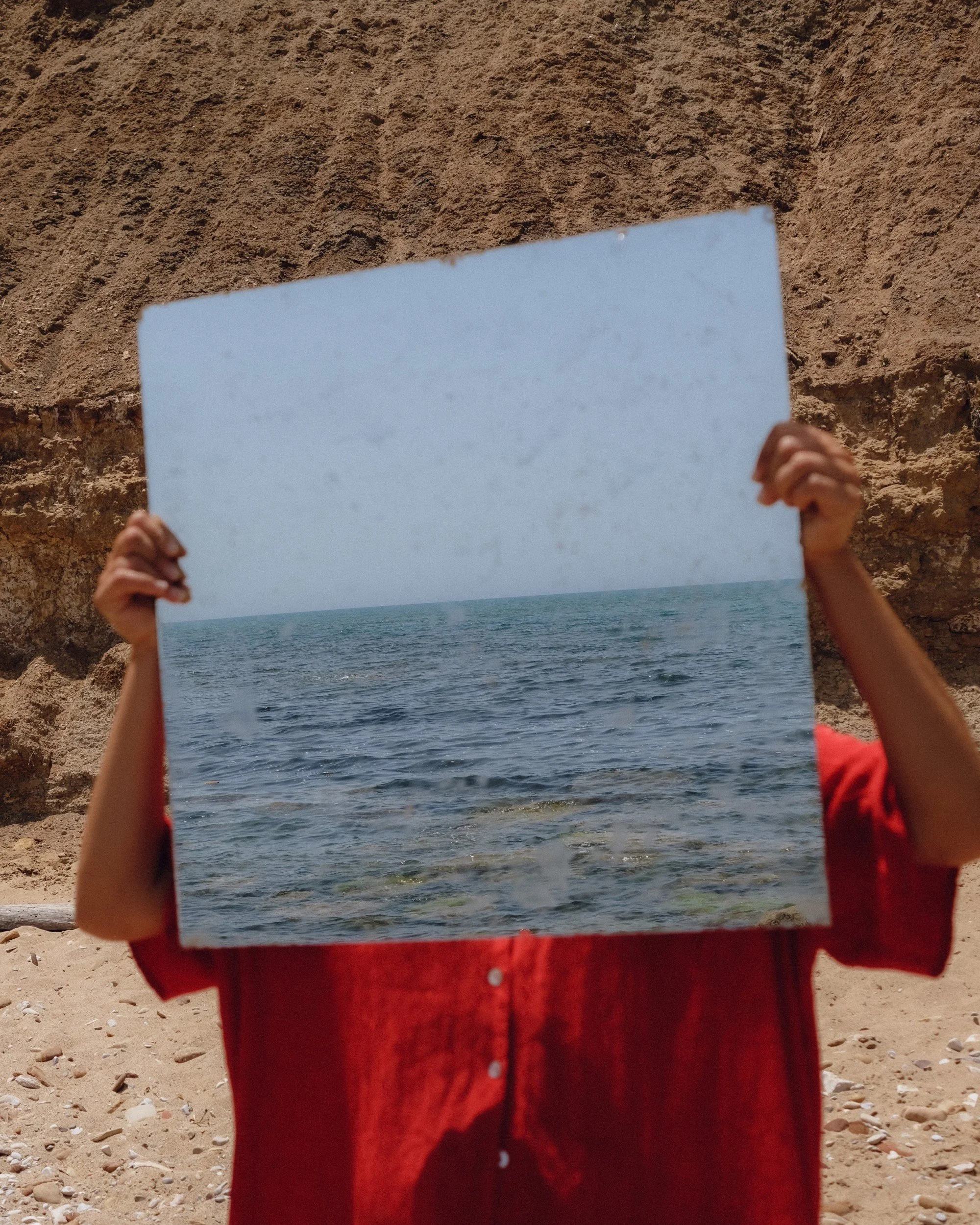 A person holding a mirror reflecting the ocean and sky with a rocky cliff in the background.