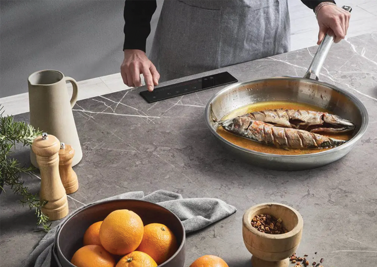 Person cooking fish in a frying pan on a gray kitchen countertop, with oranges, spices, and kitchen utensils nearby.