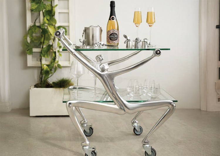 A futuristic metallic party cart on wheels with a glass top, holding a bottle of champagne, two glasses, and serving ware, set in a modern room with a potted plant.