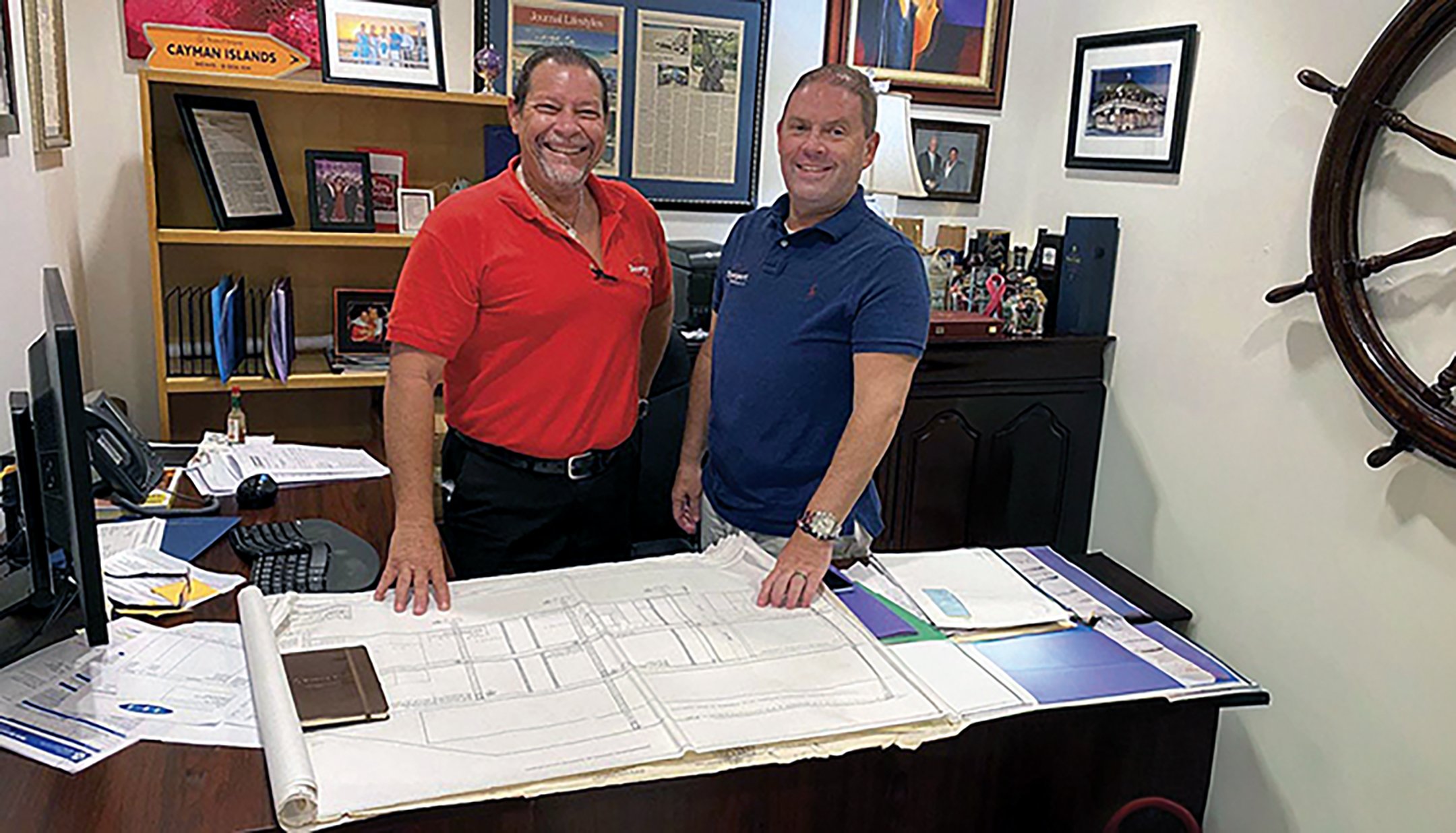 Two men standing in an office behind a desk with architectural plans. One man is wearing a red polo shirt, and the other is in a blue polo shirt. The office has framed pictures, awards, and decorations on the wall and shelves, including a ship's wheel on the right.