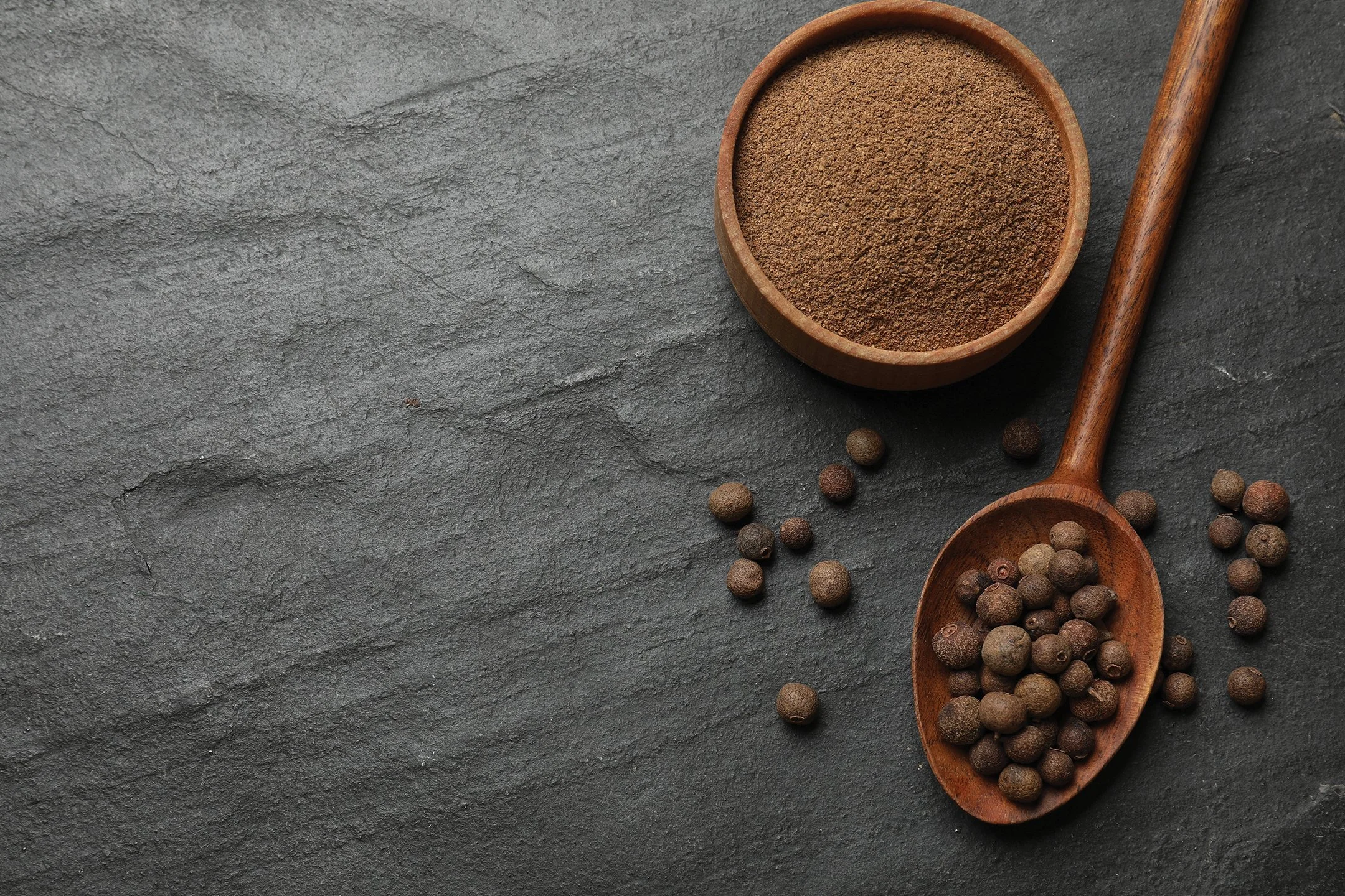 Ground black pepper in a wooden bowl and surrounding peppercorns on a dark textured surface