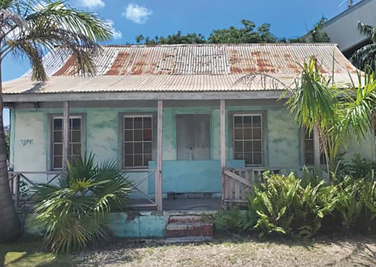 Old, weathered house with a rusty corrugated metal roof, surrounded by tropical plants, in a sunny outdoor setting.