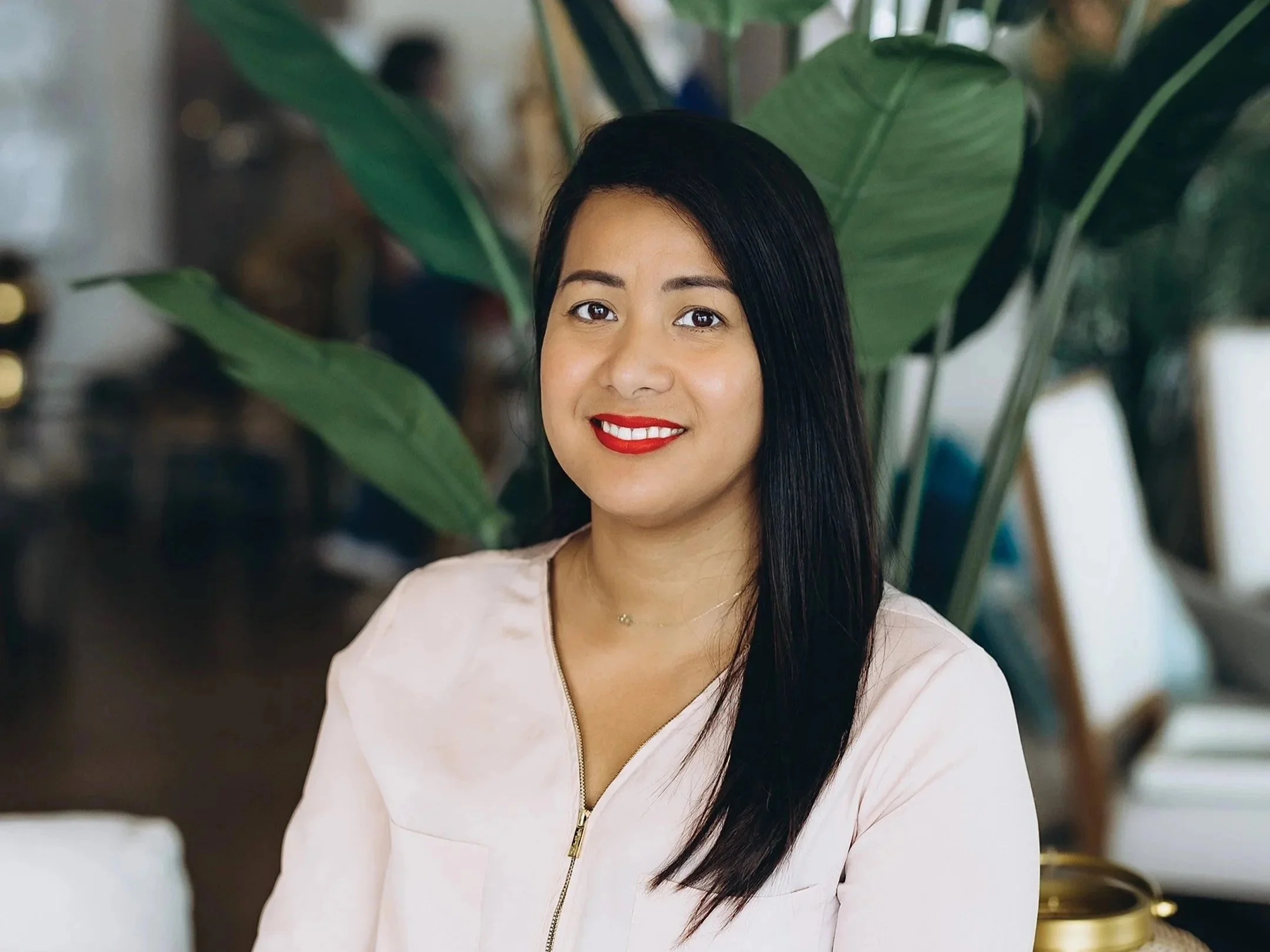A woman with straight black hair, wearing a light-colored top with a zipper, smiling and looking at the camera, in front of large green plants in an indoor setting.