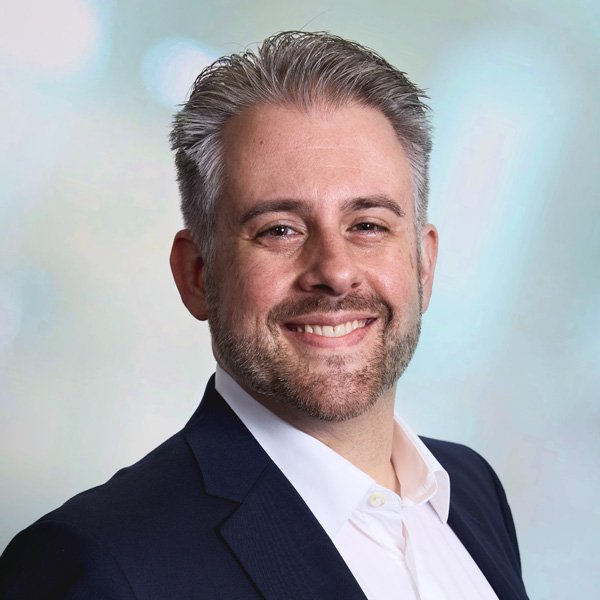 Smiling man with a beard in a suit and white shirt on a gray background.