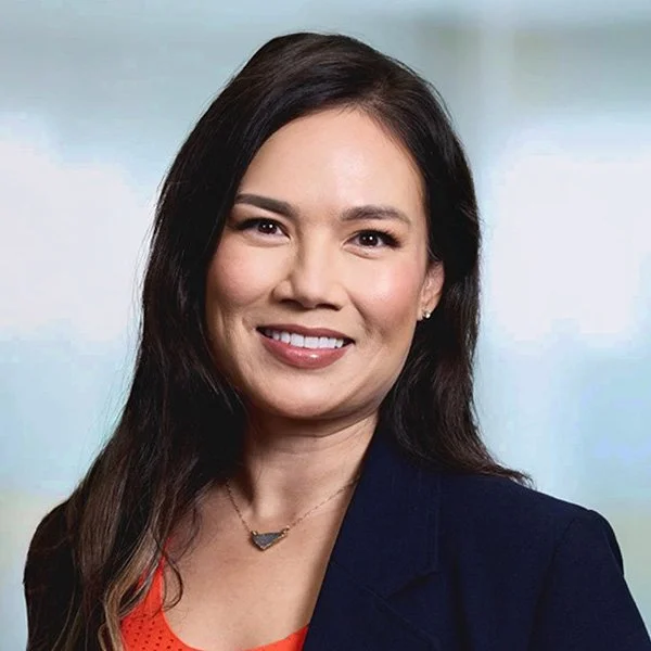Smiling woman with long dark hair, wearing a black blazer, against a gray background.