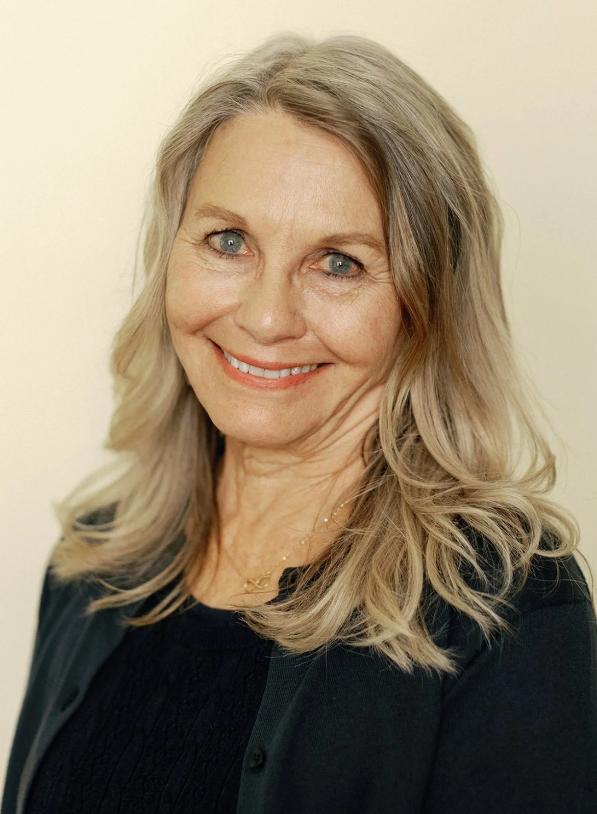 A smiling middle-aged woman with wavy blonde hair and blue eyes, wearing a black top and a gold necklace, standing against a plain light-colored background.