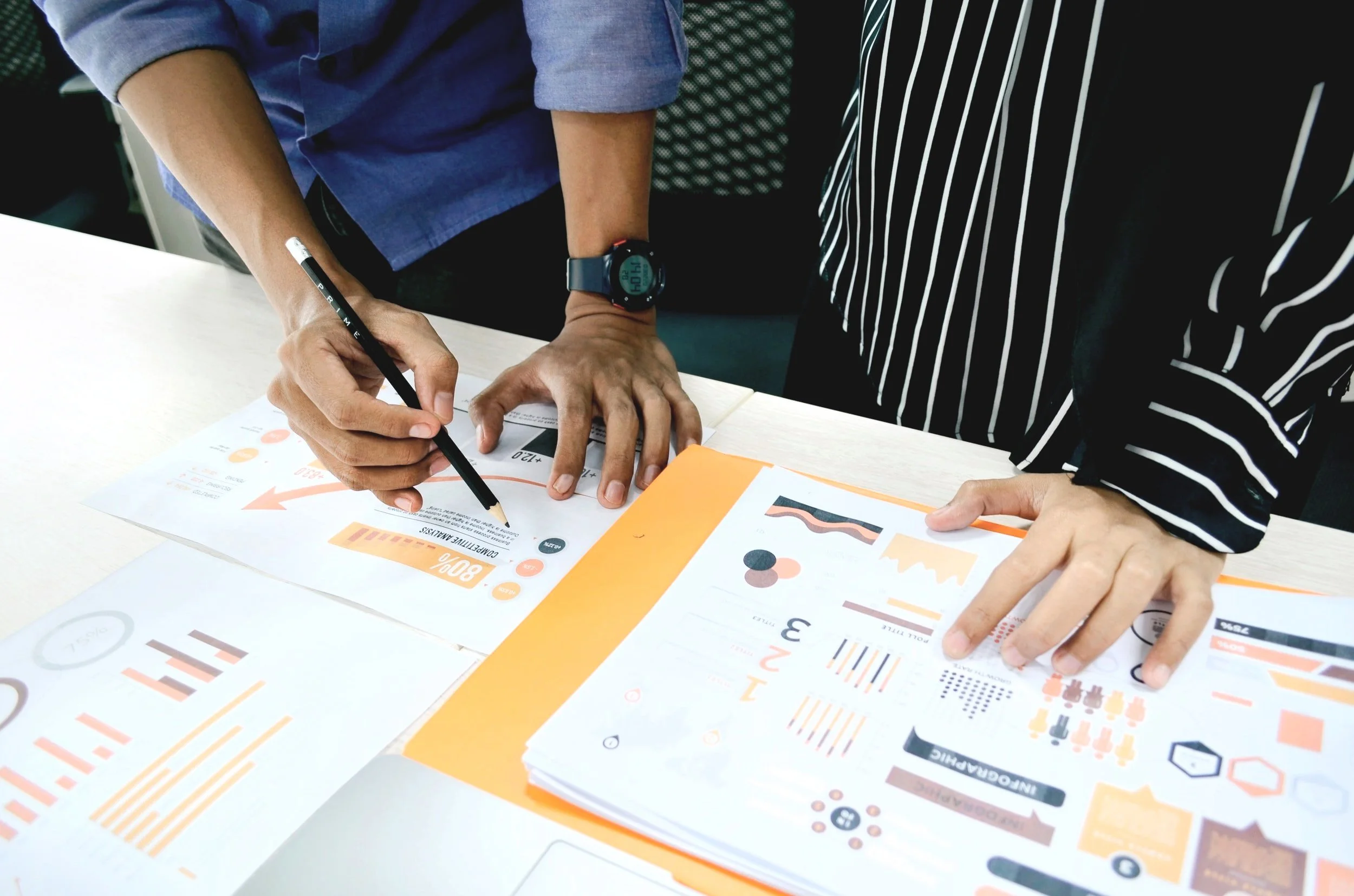 Two people working together at a desk with printed charts, graphs, and colorful infographics, one person pointing at a document while the other is holding a pen.