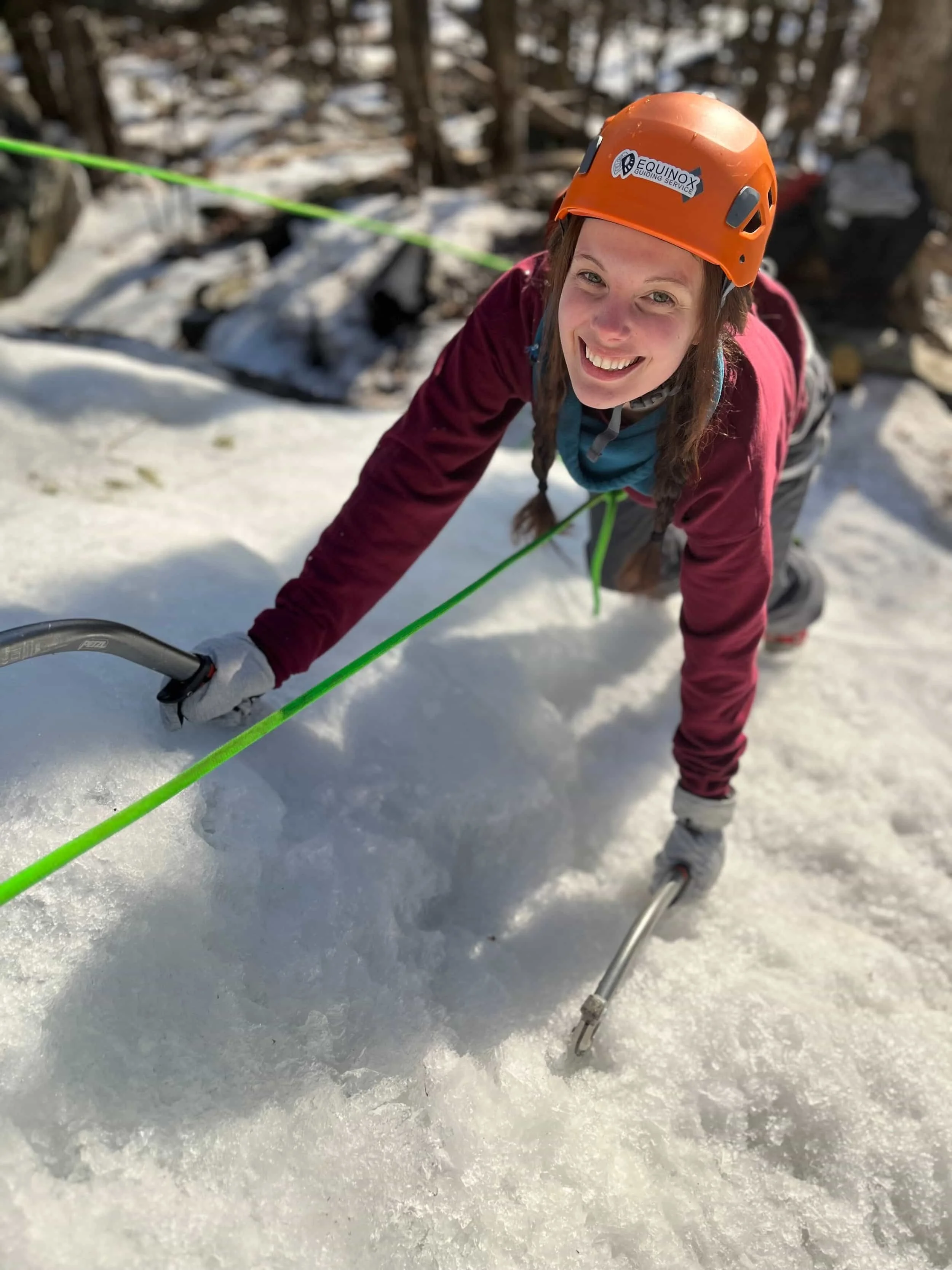 A woman with braided hair, wearing an orange helmet and a maroon jacket, is crawling on snow while holding ice axes, smiling at the camera.